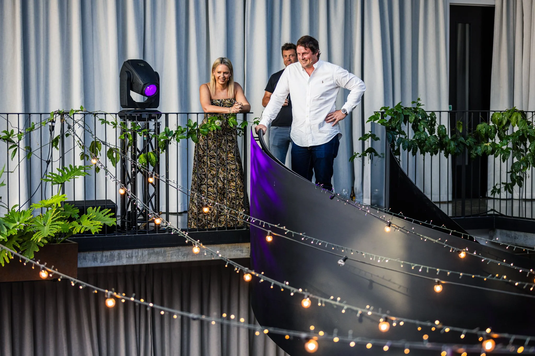 People standing on a balcony decorated with string lights and greenery, with a gray curtain in the background.