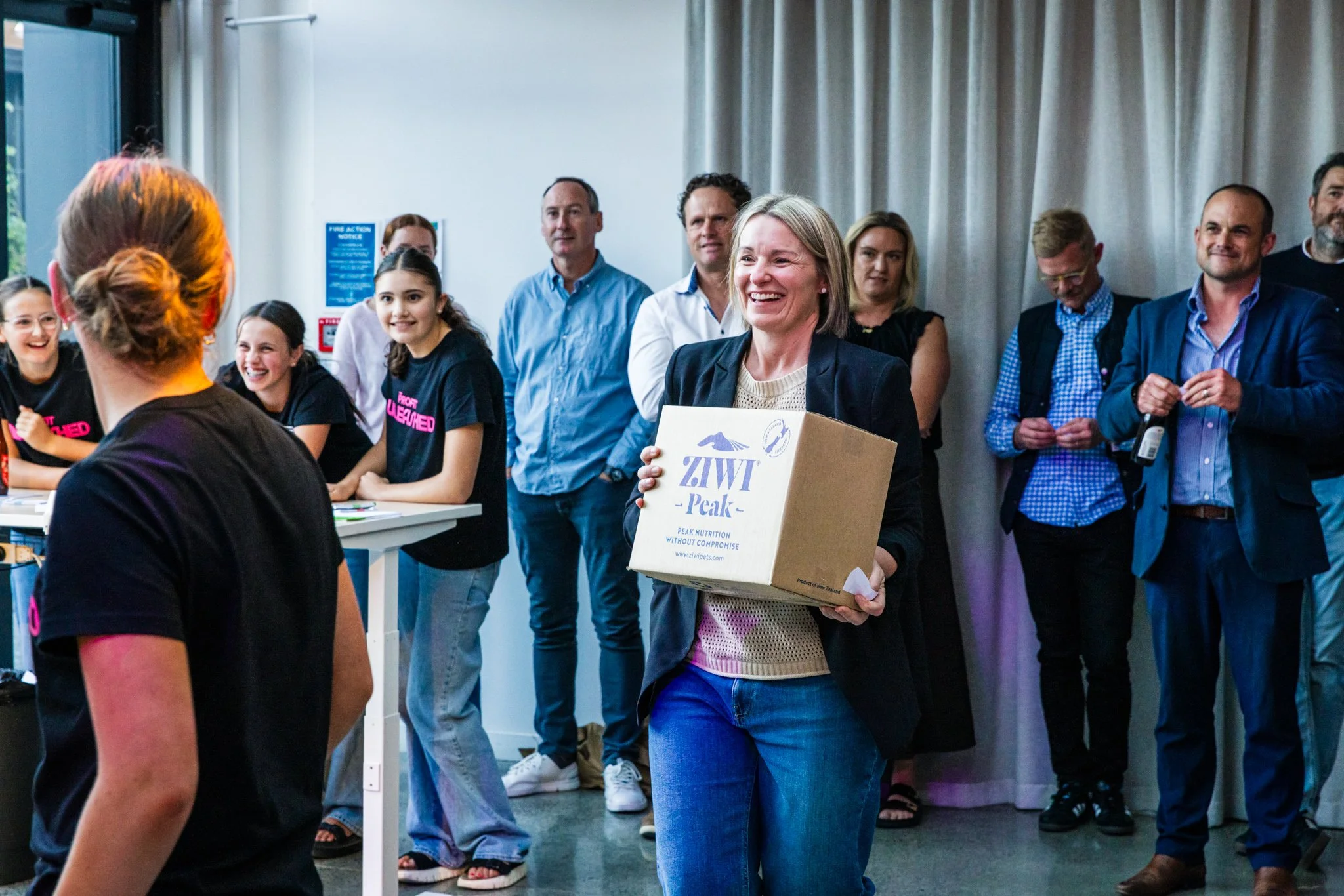 A group of people gathered indoors for a presentation or event, with a woman in the center smiling while holding a box labeled 'ZIWI Peak'.