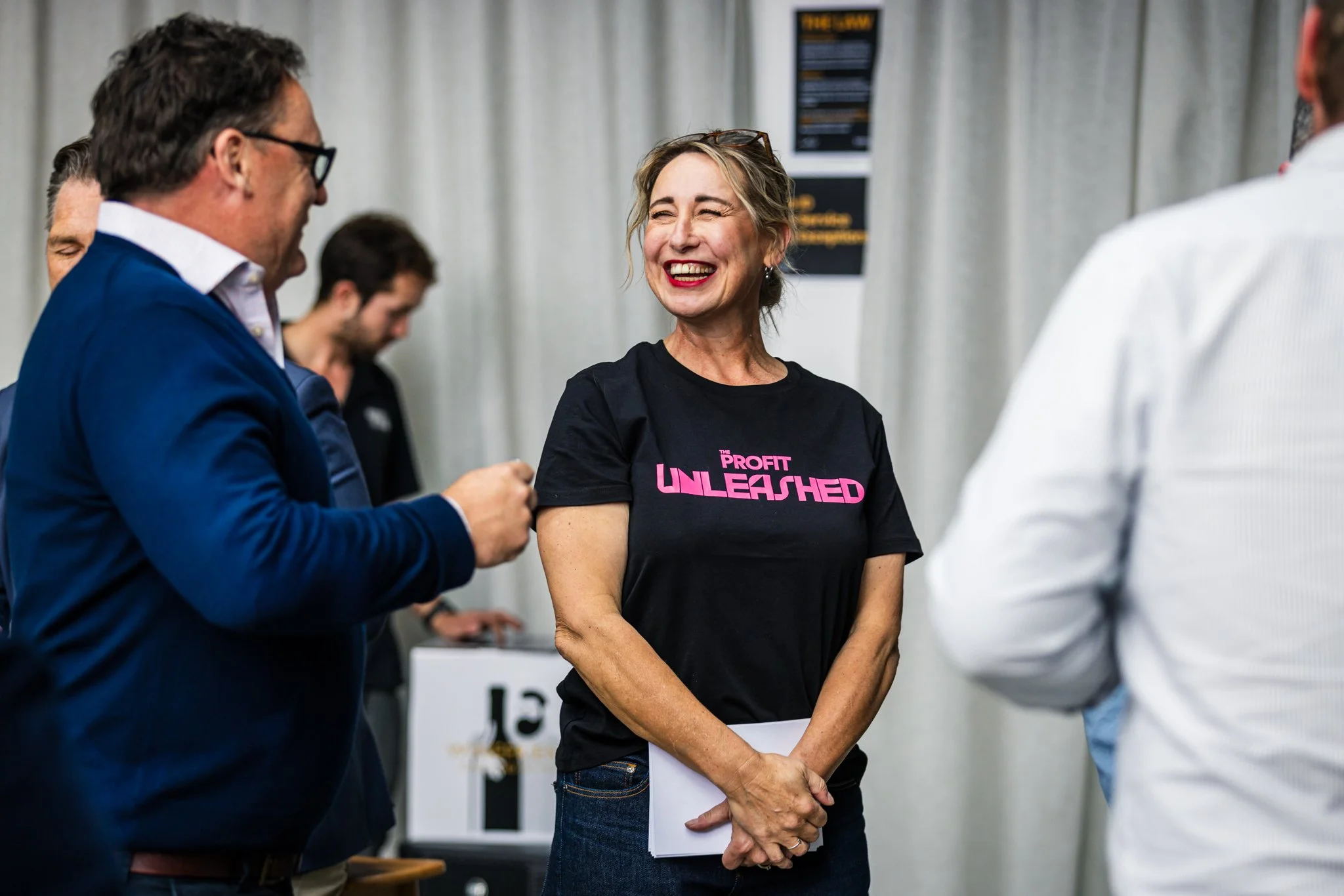 A woman wearing a black T-shirt with pink lettering that reads 'The Profit UNLEASHED' is smiling and talking with a group of people at an indoor event or conference.