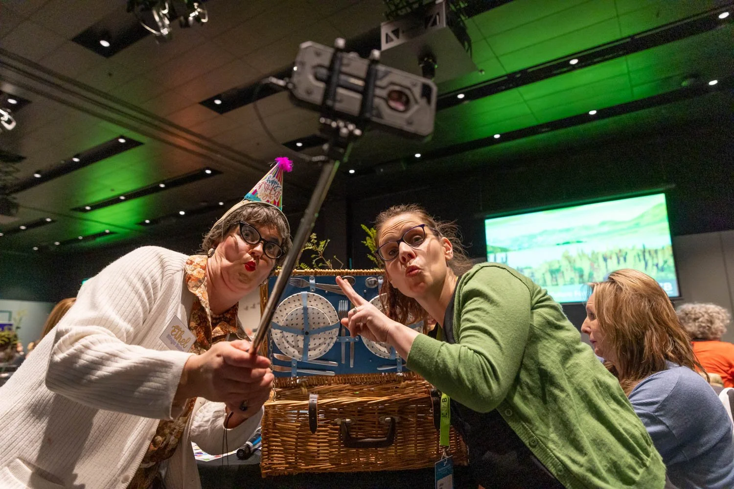 Two women taking a selfie at a celebratory event, wearing party hats, with a large computer monitor and other attendees in the background.