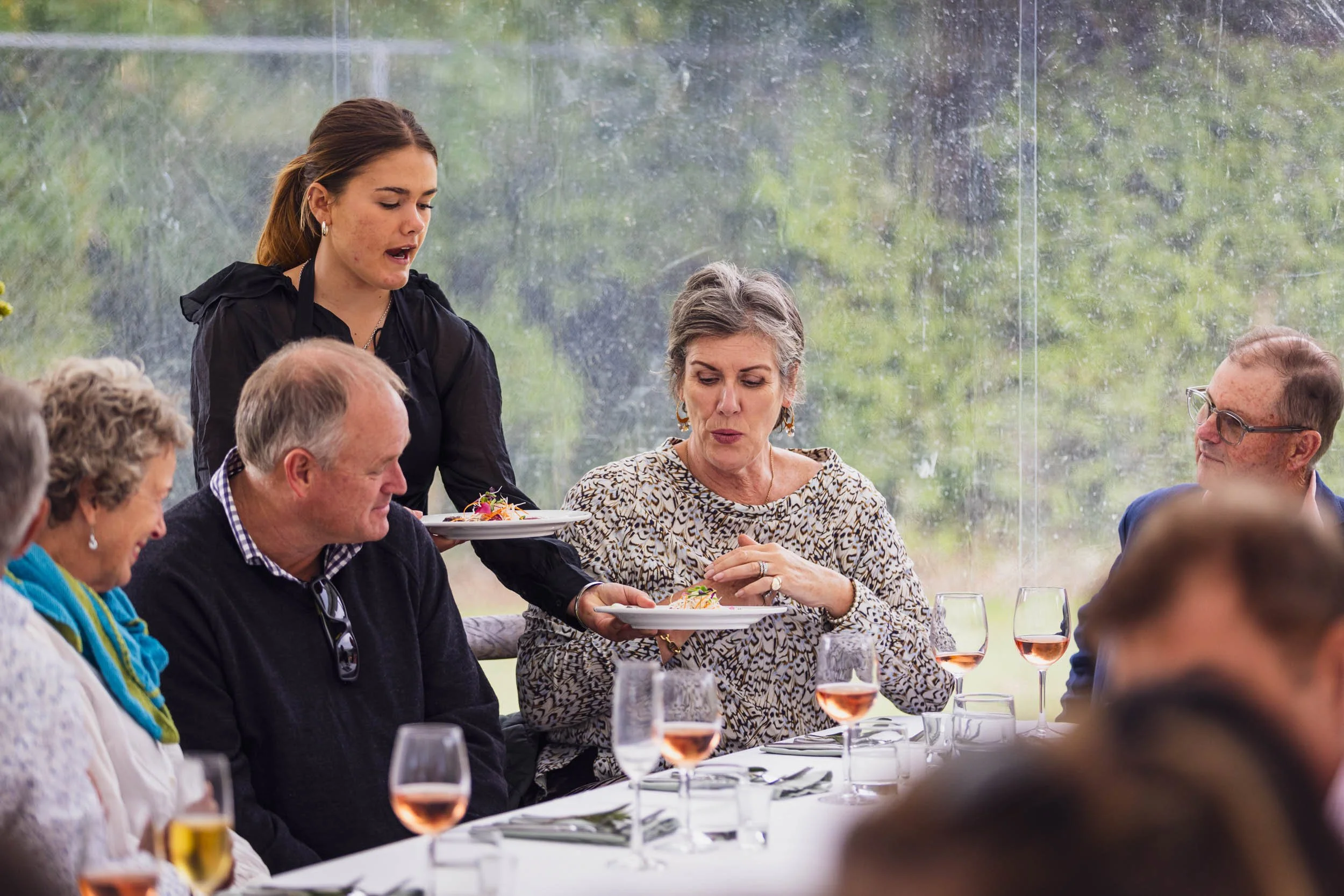 A group of people sitting at a dining table with glasses of rosé wine during a meal, with a waitress serving food, inside a glass-enclosed area with greenery outside.