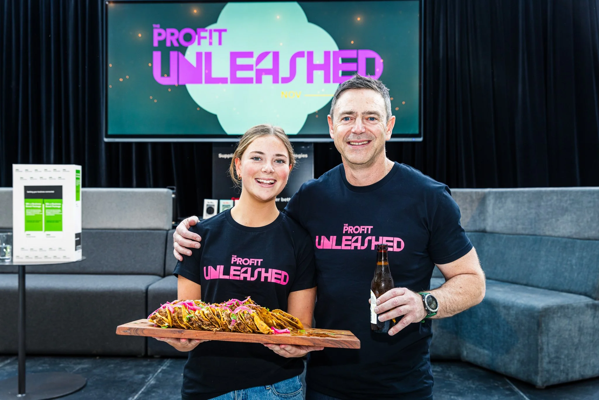 Two smiling people wearing black T-shirts with pink text that reads "The Profit Unleashed" standing together in a room with black curtains, holding a wooden tray of tacos and a bottle of beer, with a large screen behind them displaying the same text.