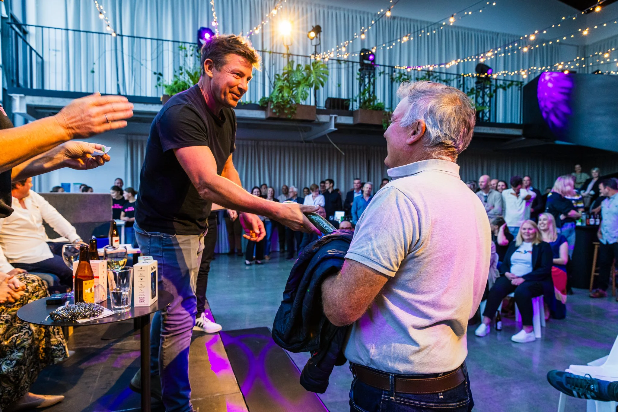 A man in a black T-shirt shakes hands with an older man in a white polo shirt at a lively indoor event with string lights and a crowd of people seated and standing.