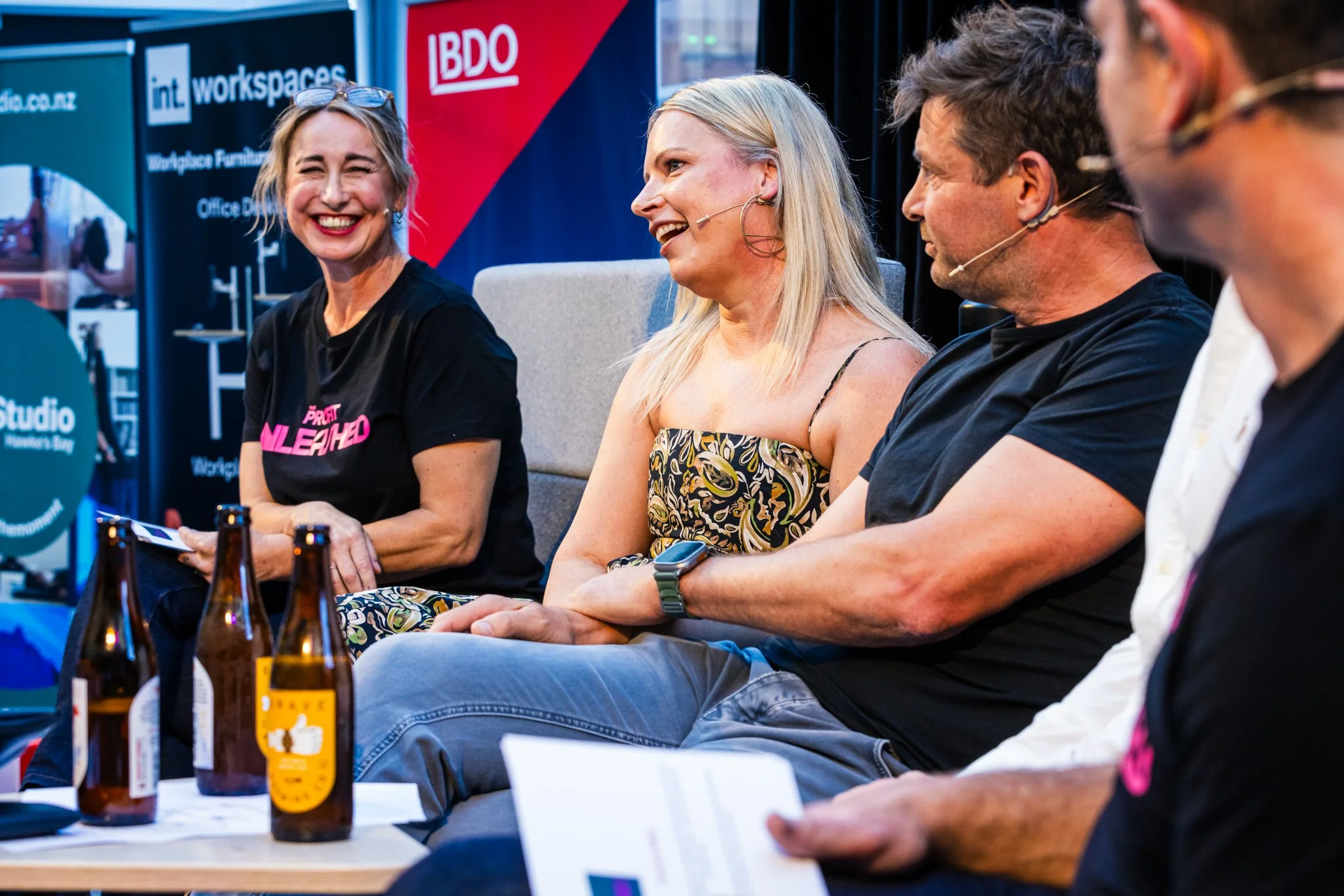 Four people sitting on stage at a panel discussion, with a woman in a black T-shirt smiling, and others engaging in conversation, behind a table with three beer bottles.