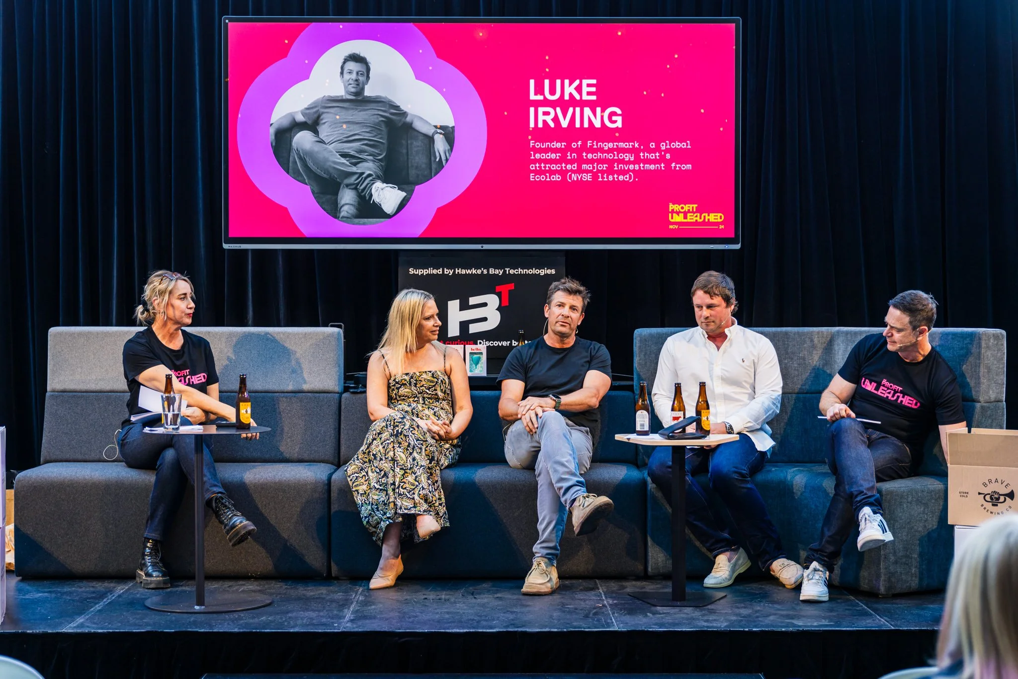 Panel discussion with five people sitting on stage, three men and two women, with drinks and a microphone, in front of a large screen displaying information about Luke Irving, founder of Fingermark, and a black backdrop.