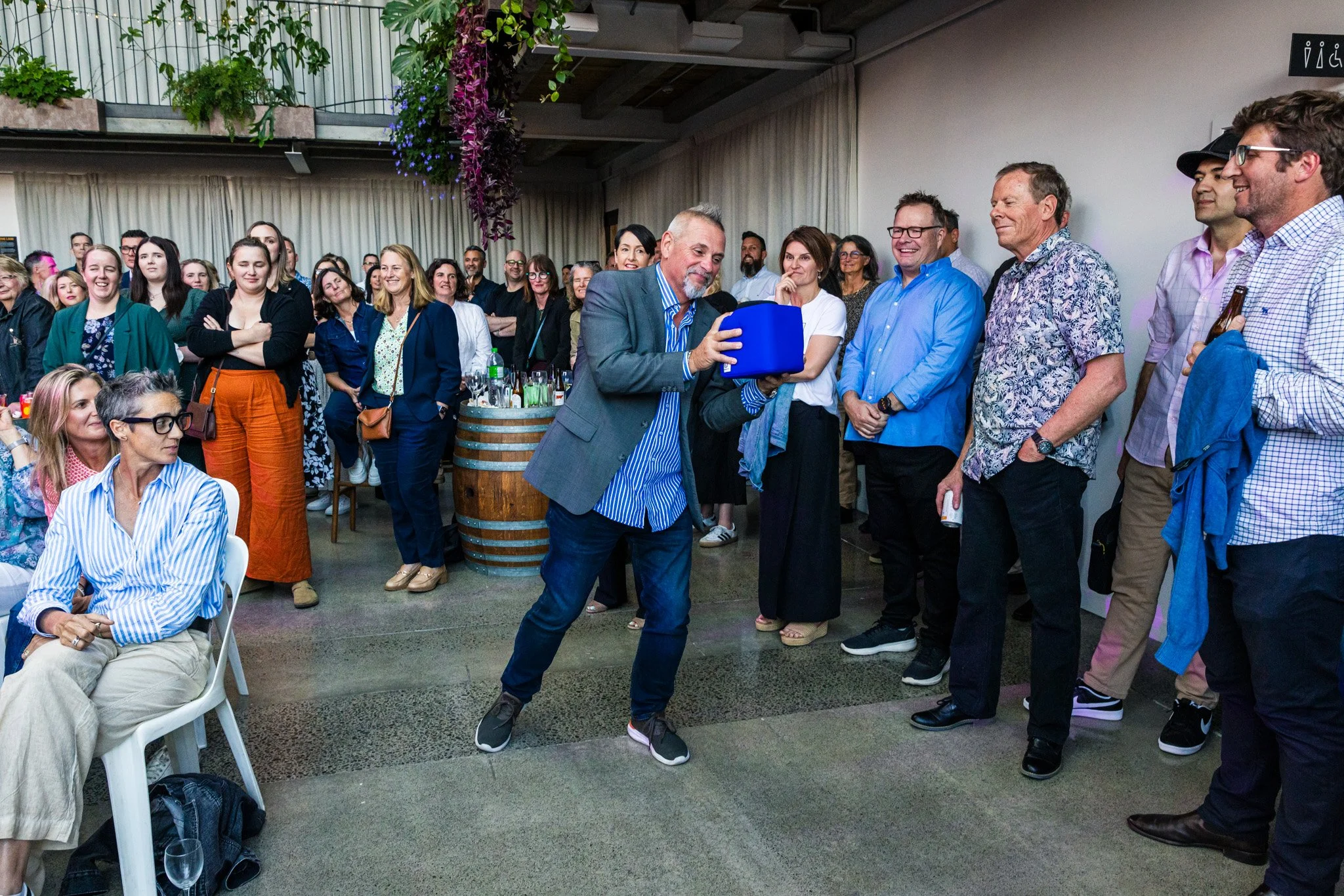 A man in a gray blazer and striped shirt is opening a blue box at a gathering of people indoors, with some people smiling and watching while others are standing and sitting.