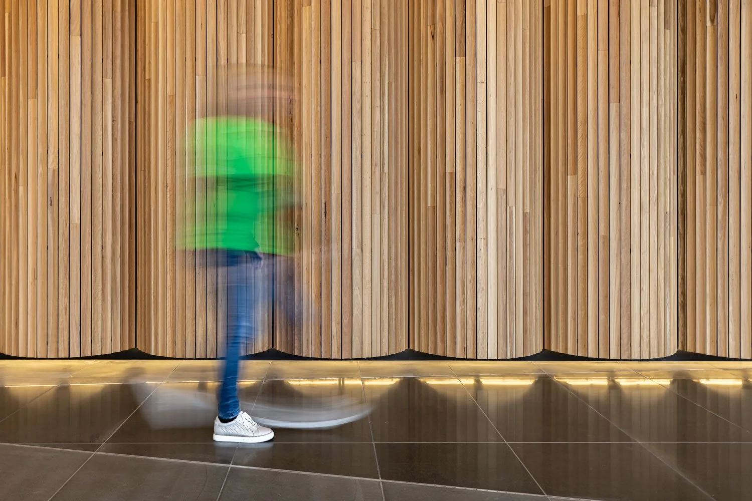 Person walking past a wooden wall with motion blur, wearing a bright green top, blue jeans, and white sneakers.
