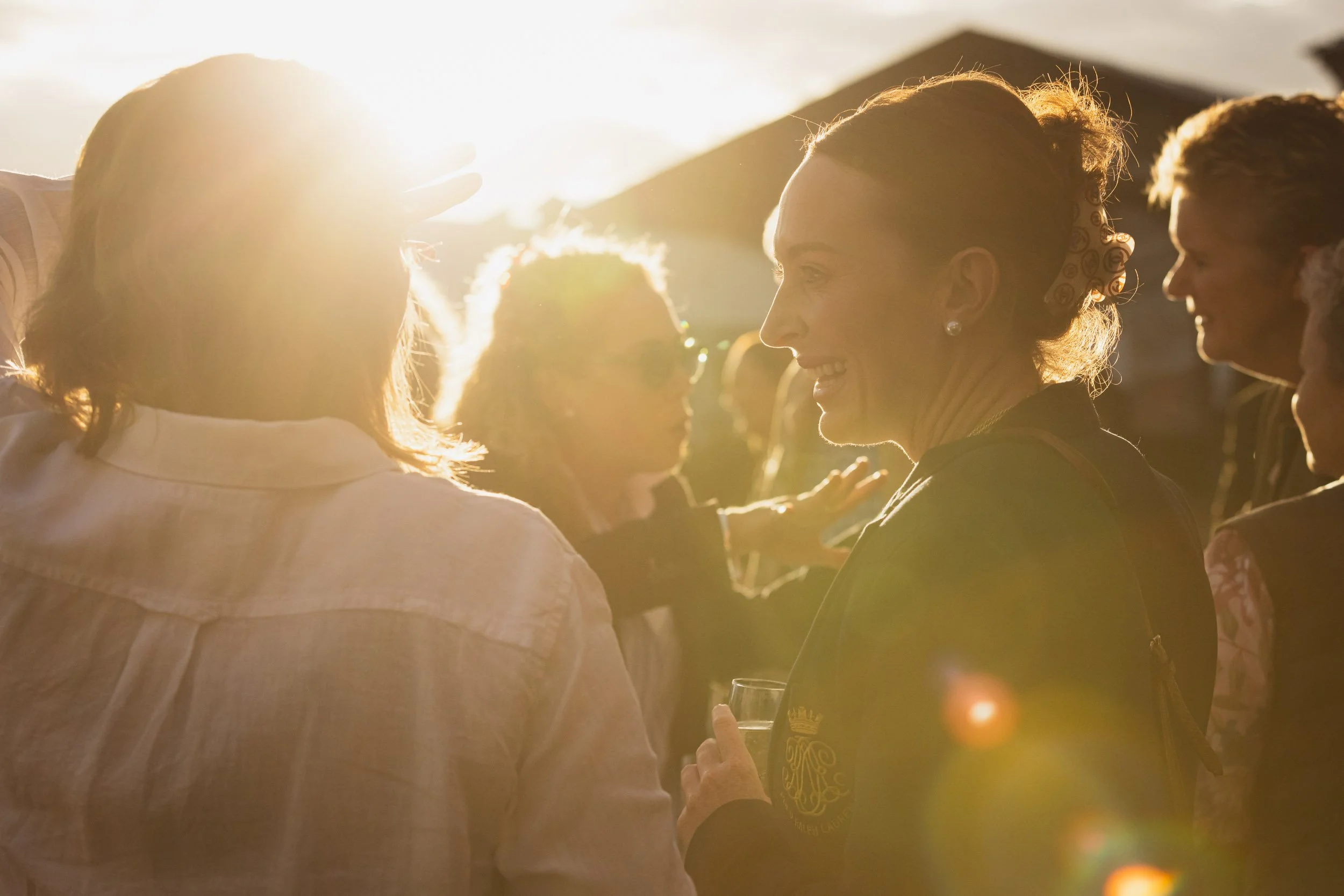Group of women socializing outdoors in sunlight, smiling and holding drinks.