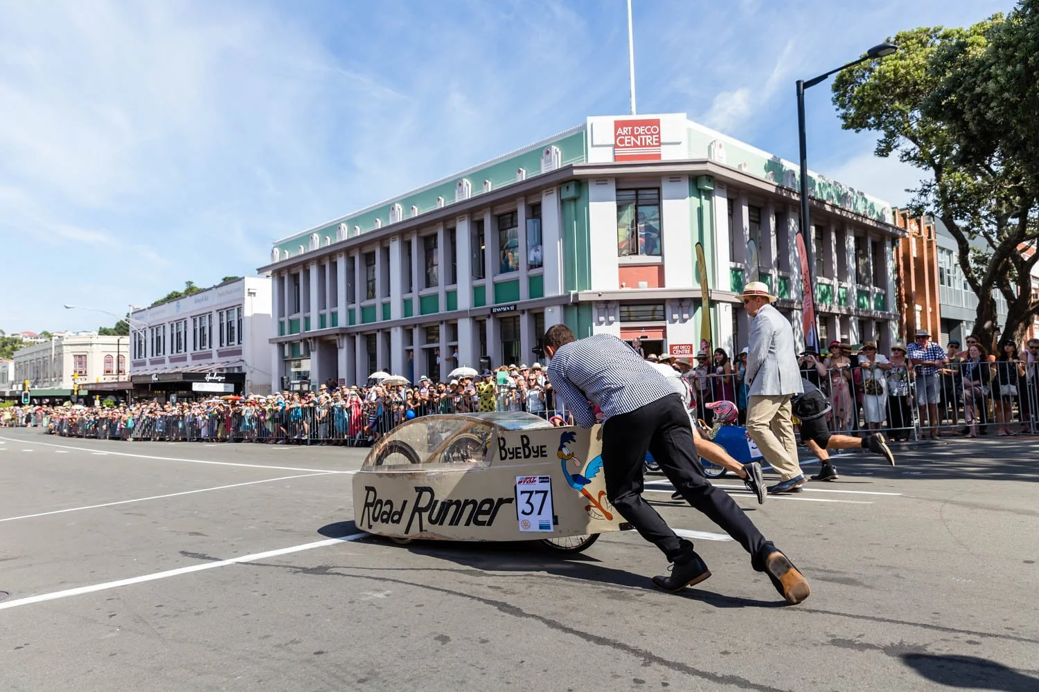 People pushing a solar-powered car during a race or parade on a city street with a crowd behind barriers and a large building with signs, including Art Deco Centre, in the background.