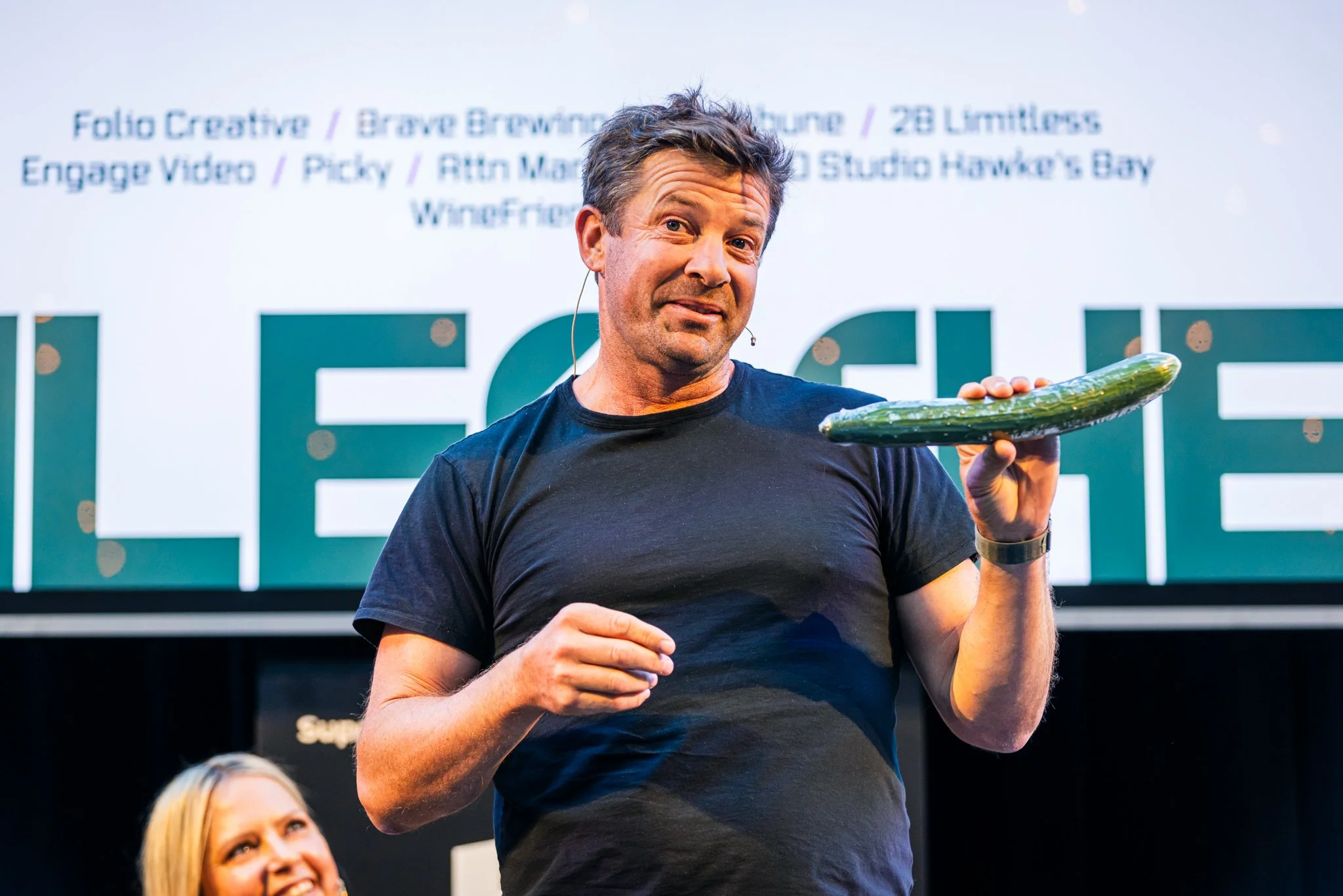 A man holding a cucumber on stage during a presentation with a woman smiling in the background.