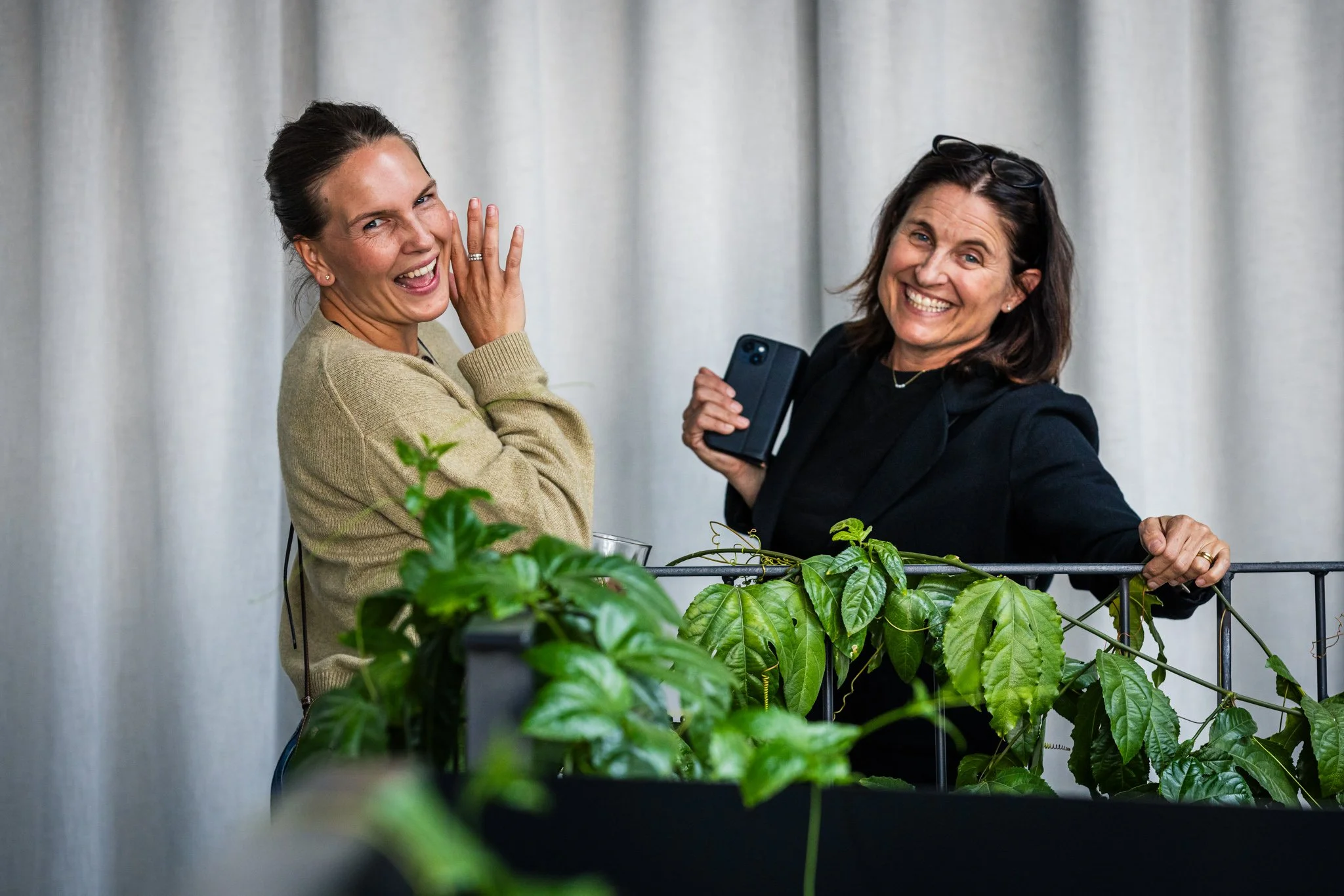 Two smiling women in front of a backdrop of greenery, taking a selfie together, one with a smartphone, both in casual and professional attire.