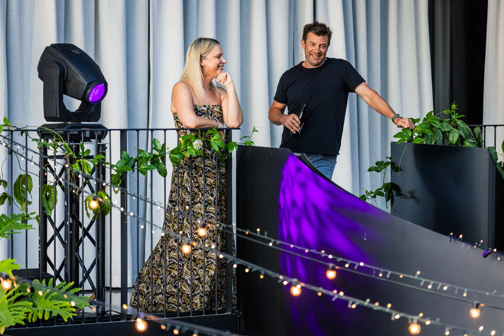 A woman and a man standing on a balcony with plants and string lights, smiling and talking, during a social event.
