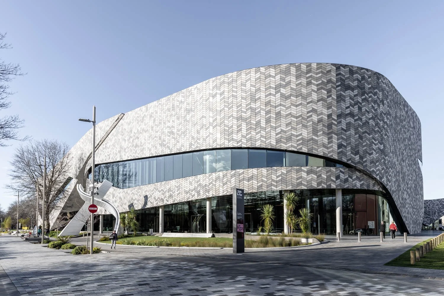 Modern museum building with a curved gray patterned facade, large glass windows, and an outdoor staircase, surrounded by trees and a paved walkway.