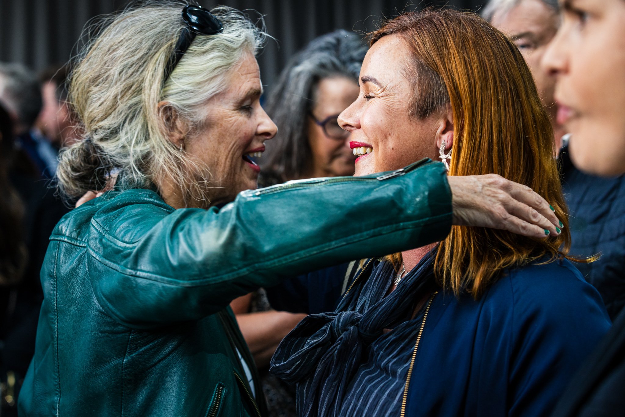 Two smiling women embrace warmly in a crowd, the woman on the left has gray hair and wears a teal leather jacket, and the woman on the right has red hair and wears a dark jacket with a scarf.
