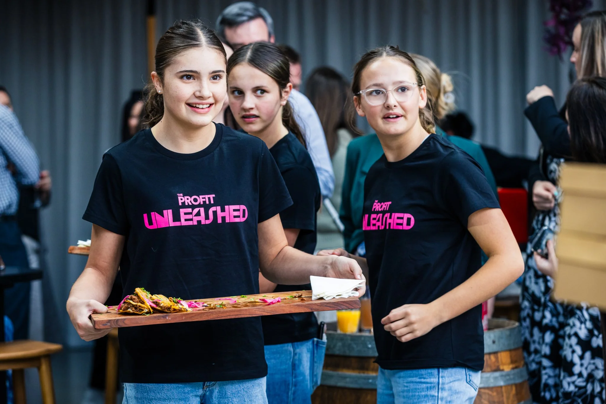 Two young girls wearing black T-shirts with pink text that reads 'Profit Unleashed' stand in a lively indoor event. The girl on the left holds a wooden serving tray with food, and the girl on the right looks to the side. Several other people are in t