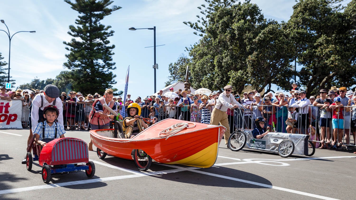 Children in vintage-style racing carts at a parade, with people watching behind a barrier, some holding umbrellas, under a partly cloudy sky and surrounded by trees.