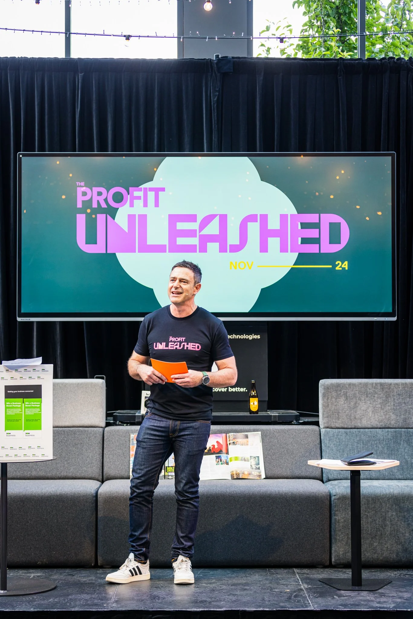 A man is standing on stage during a presentation, wearing a black t-shirt with pink text that says 'Profit Unleashed'. Behind him, a large screen displays the same phrase in bold pink letters with the date November 24. The stage is set with gray seat