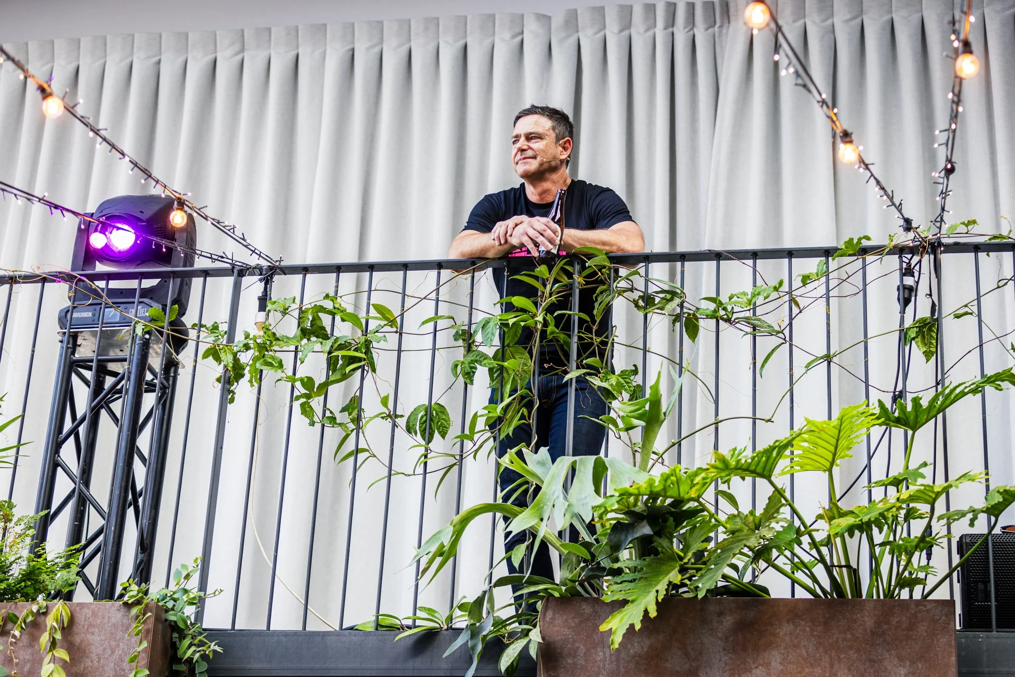 A man leaning on a balcony railing with plants in front, holding a drink, amid decorative string lights, in front of a white curtain.