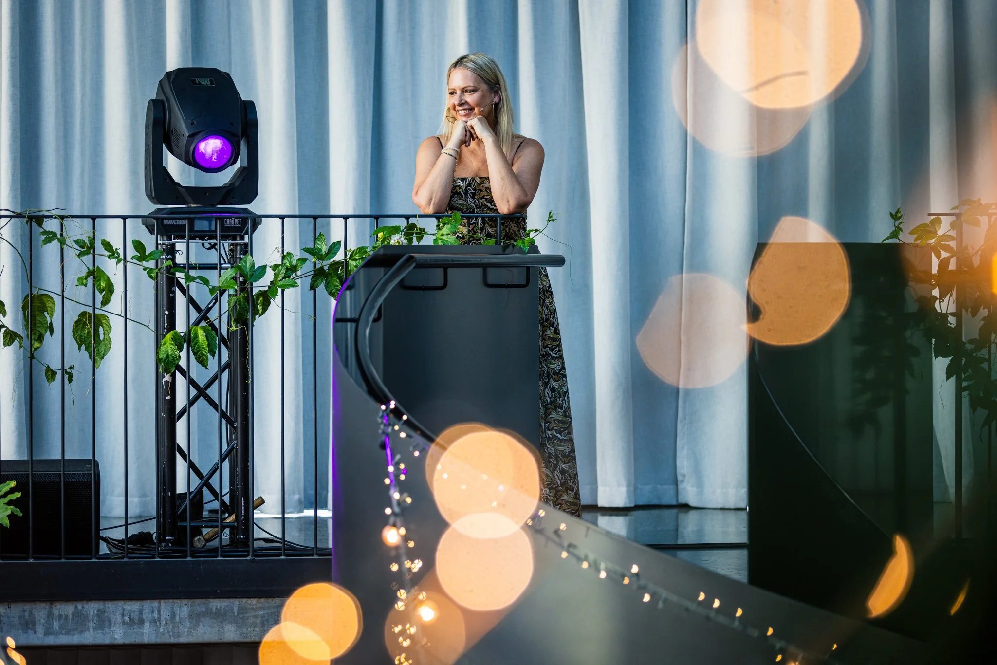 A woman in a floral dress stands on a stage, smiling with hands near her face, next to a DJ booth with plants and stage lighting.