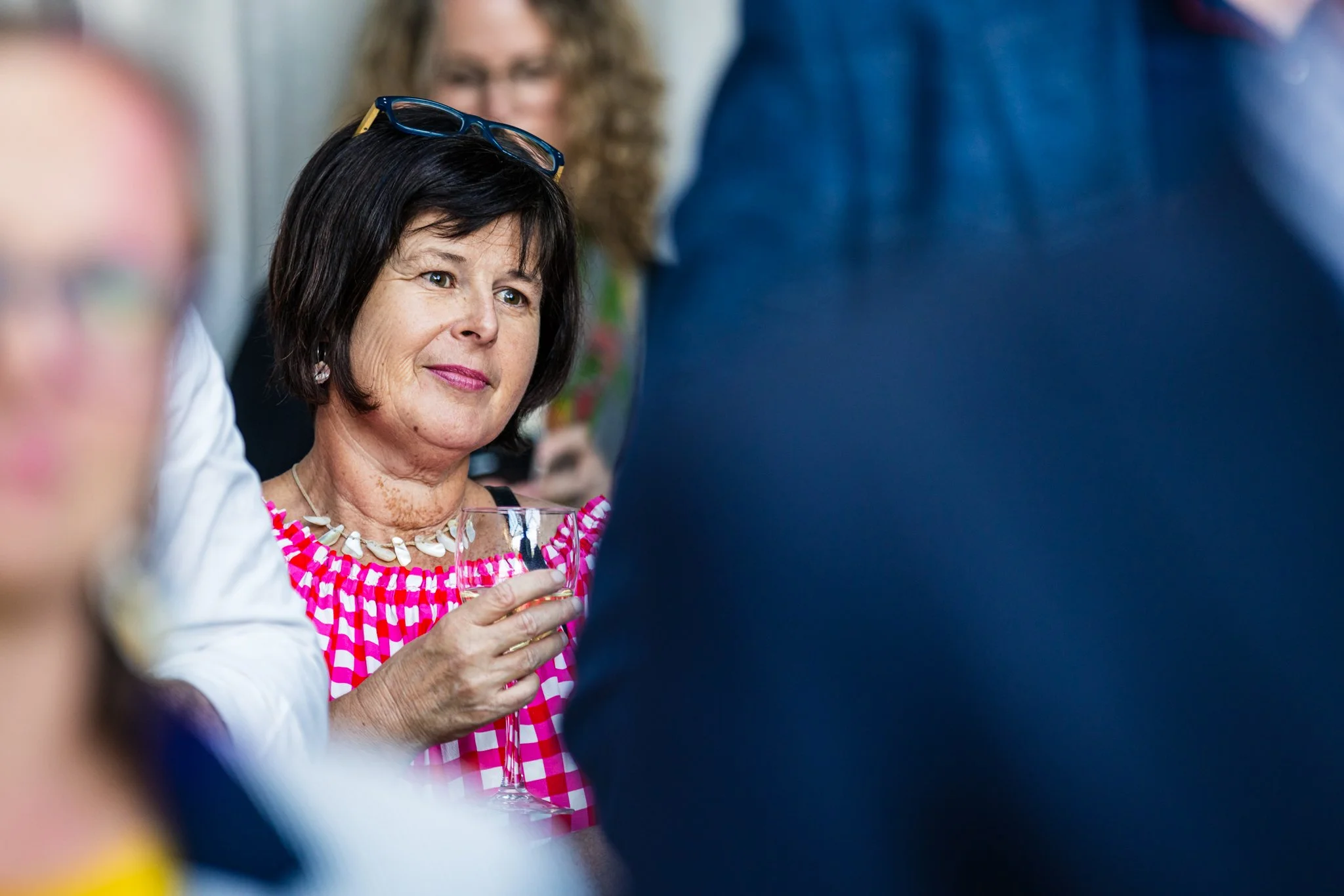 A woman with short dark hair, wearing a pink checkered top, holding a wine glass at a social gathering, with glasses, other people in the background, and blurred figures in the foreground.