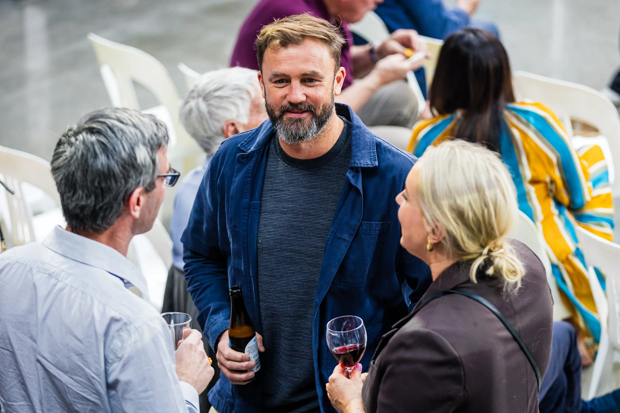 People at a social gathering talking and holding drinks, including a man with a beard in a blue jacket smiling and engaging in conversation.