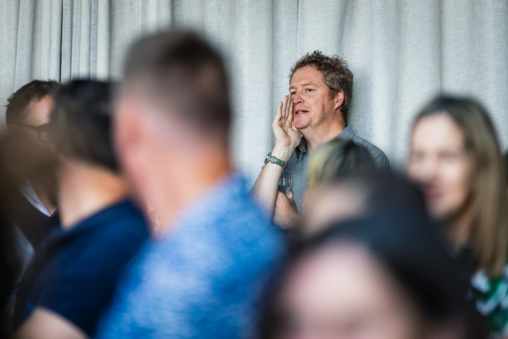 A man with curly hair and a checkered shirt is listening to someone speaking, with his hand on his cheek, in a setting with others around him.