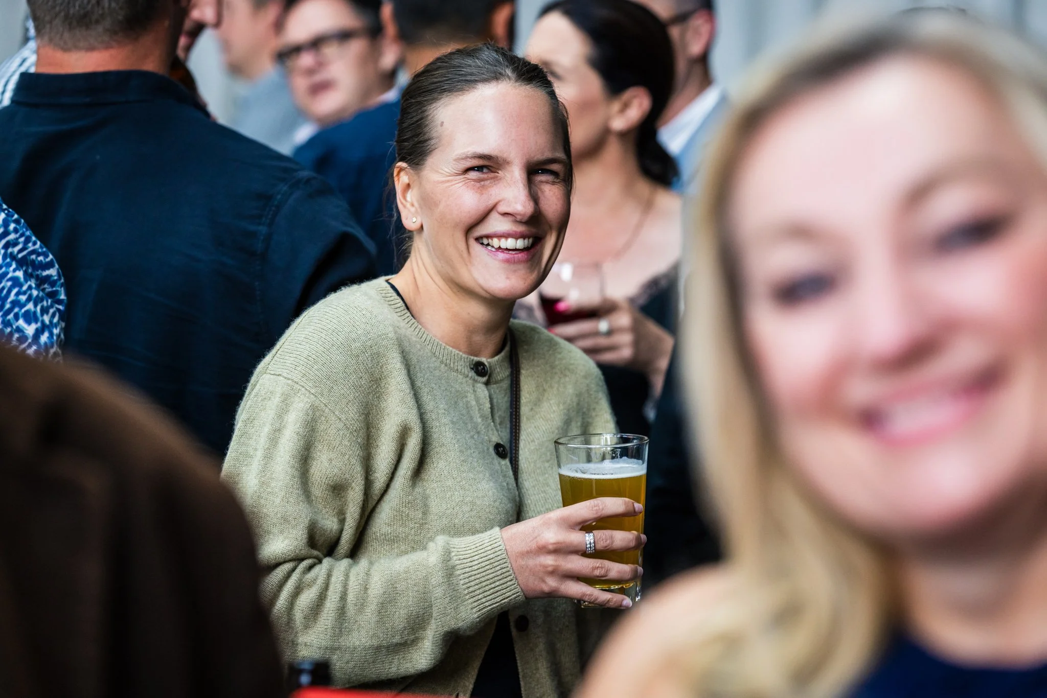 A woman with brown hair holding a glass of beer, smiling at a social gathering with other people in the background.