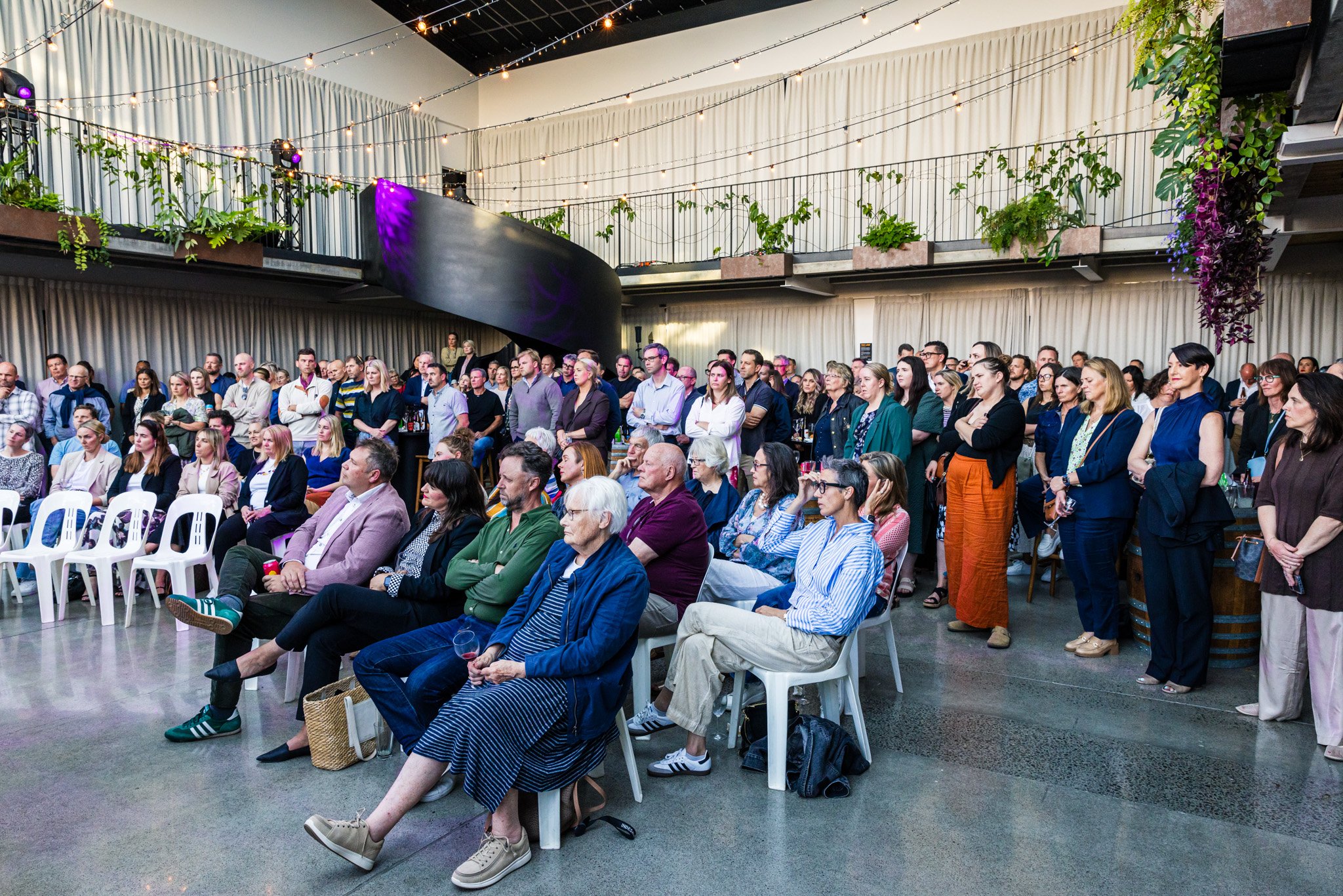 A large crowd of people attending an indoor event, seated and standing, with purple and white lighting, plants, and curtains in the background.