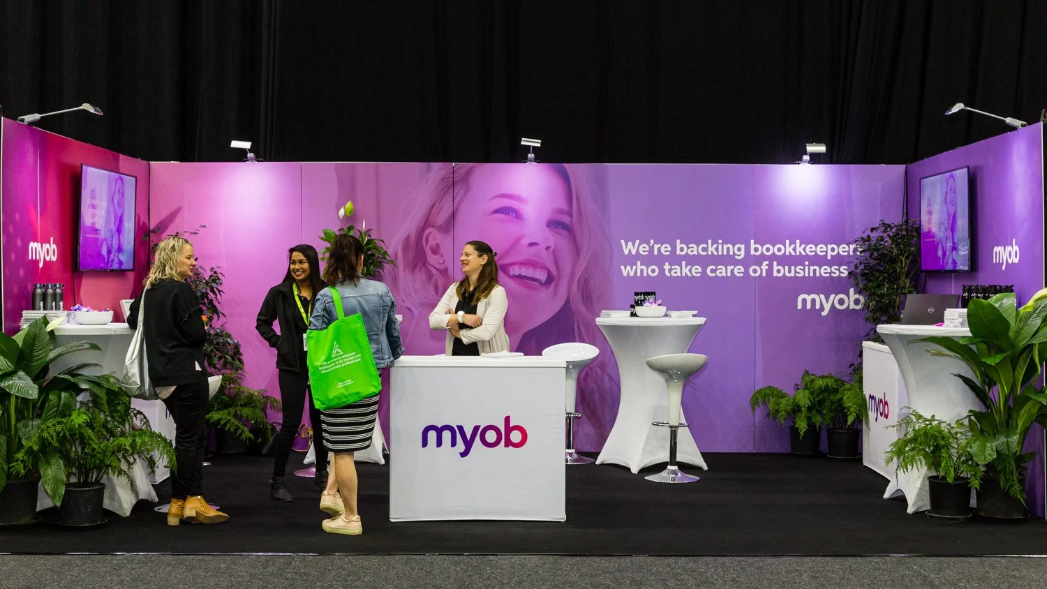 Trade show booth for myob with four women talking, plants, TV screens. Purple backdrop with smiling woman and white text reading, 'We're backing bookkeepers who take care of business.'