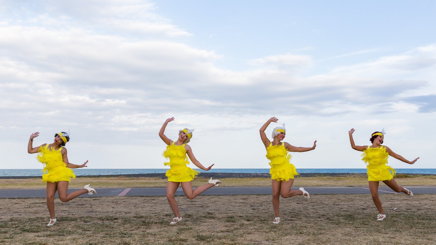 Four women dressed in yellow feathered dresses and white heels dancing on a grassy area near the beach with the ocean and cloudy sky in the background.