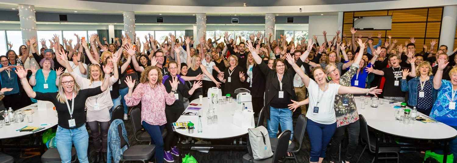 A large group of people in a conference room are raising their hands and smiling, with tables and conference materials in front of them.