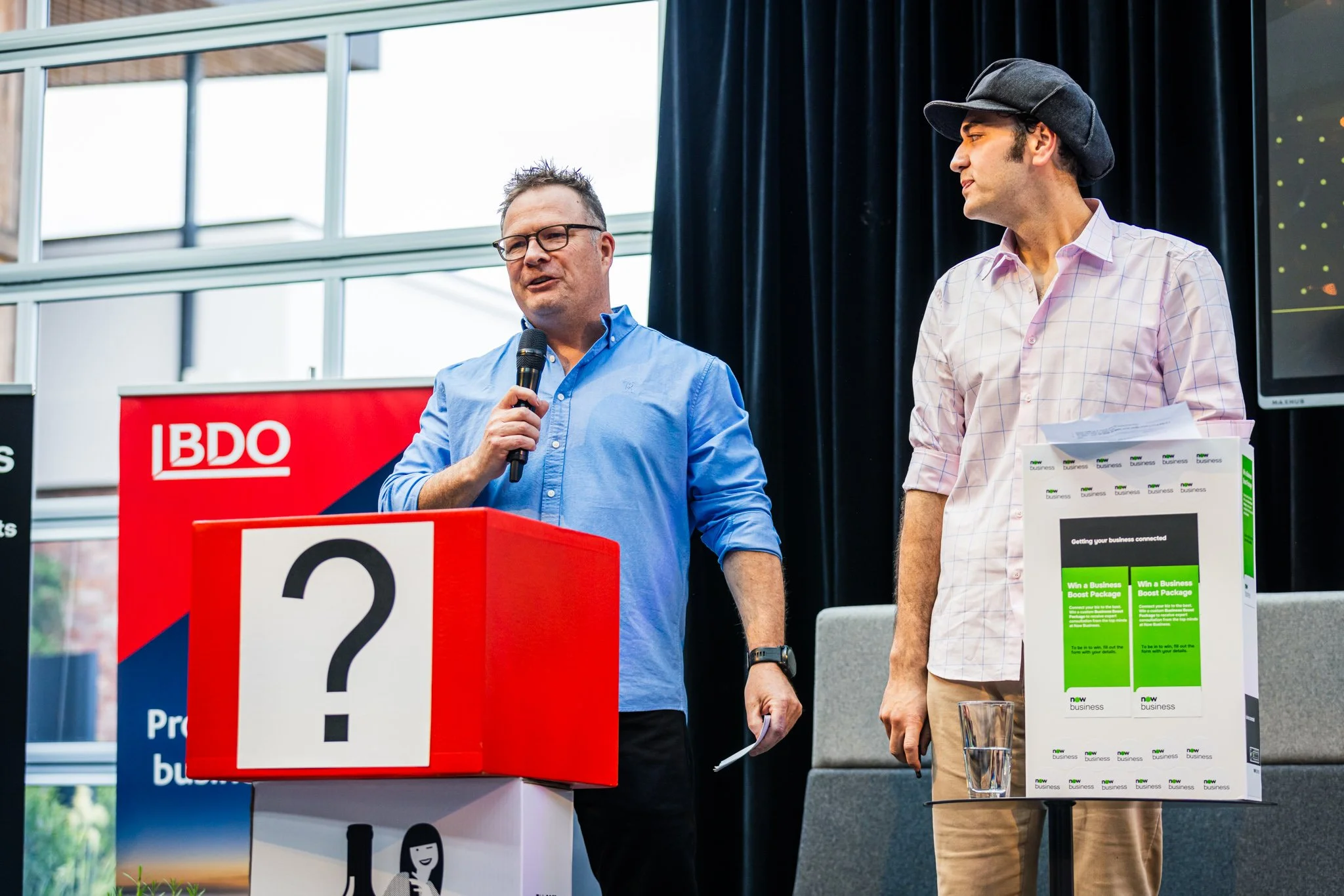 Two men standing at a presentation or event, one speaking into a microphone and the other holding papers, with a large red box with a question mark on it, and promotional materials on a stand.