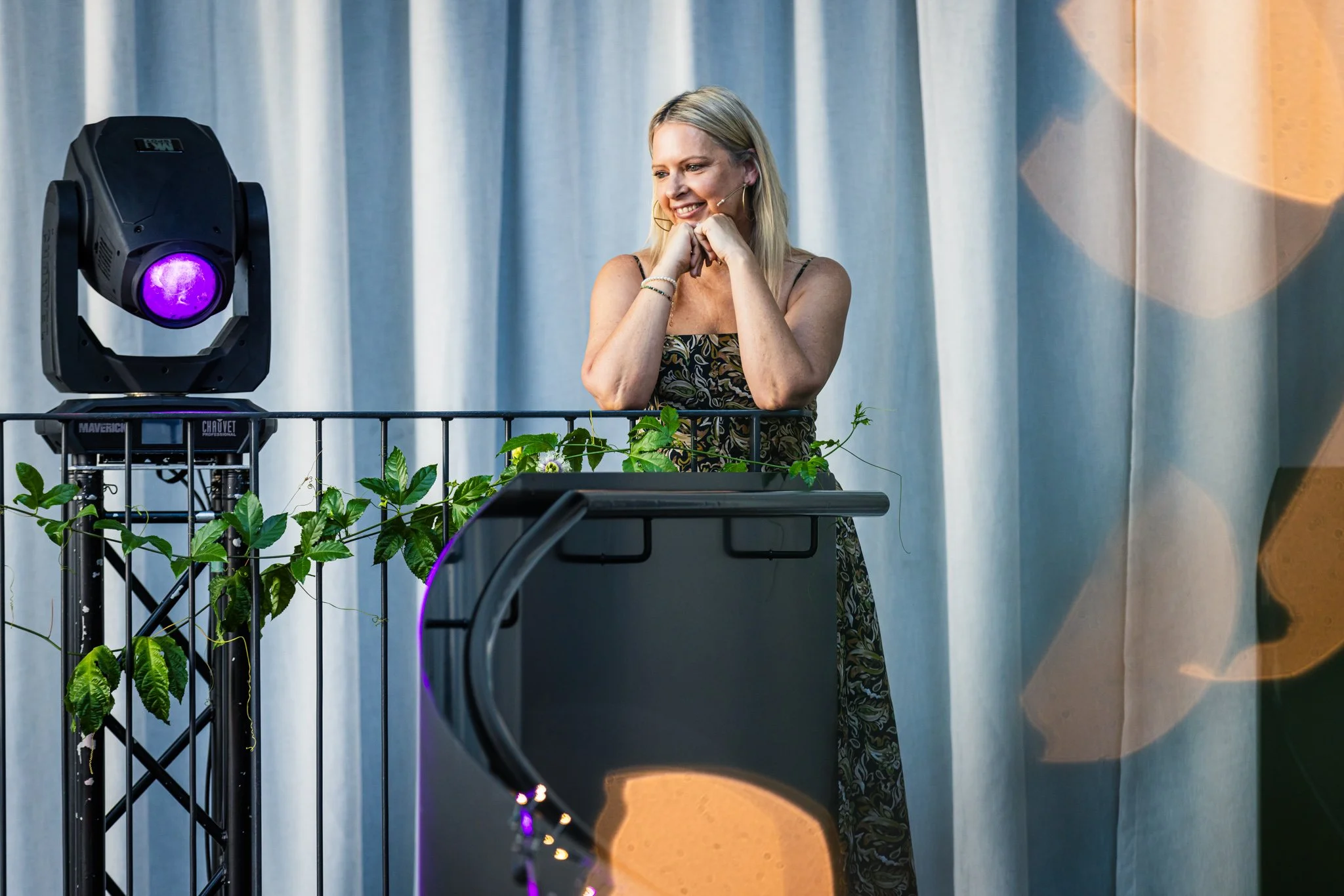 A woman with blonde hair smiling and wearing a sleeveless patterned dress stands behind a podium with a portable railing. There are green plants and stage lights around her, and a light curtain backdrop.