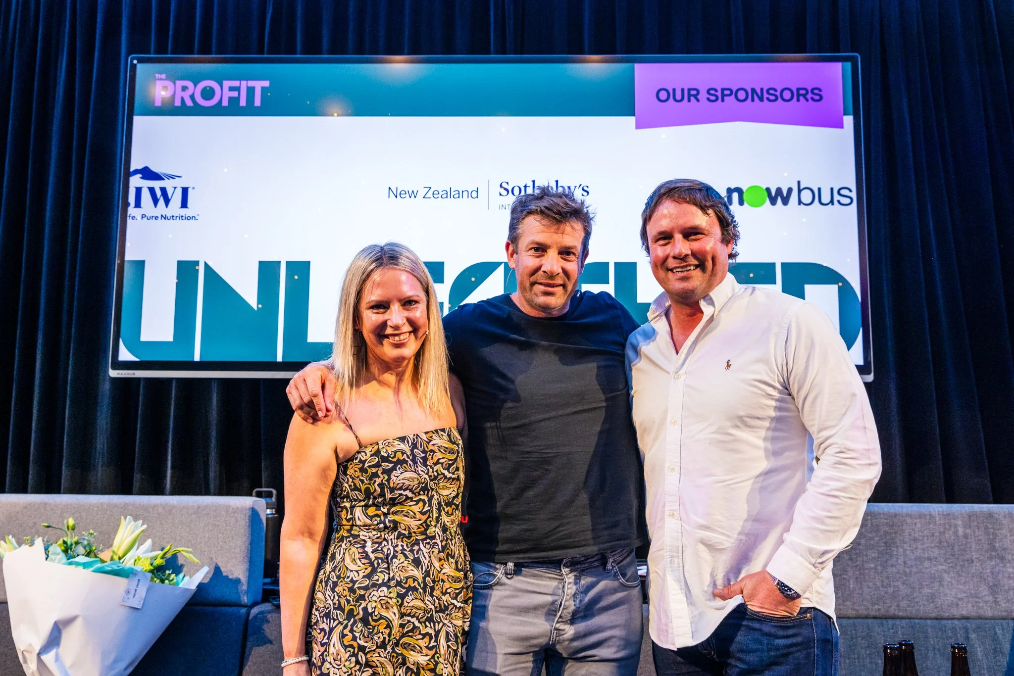 Three people standing together on stage at a conference or event, smiling at the camera. Behind them is a large screen displaying logos and the words 'Unleashed' and 'Our Sponsors.'