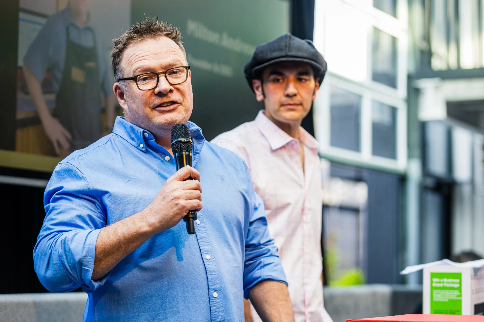 A man in a blue shirt speaking into a microphone, standing next to another man in a light pink shirt and hat at an indoor event.