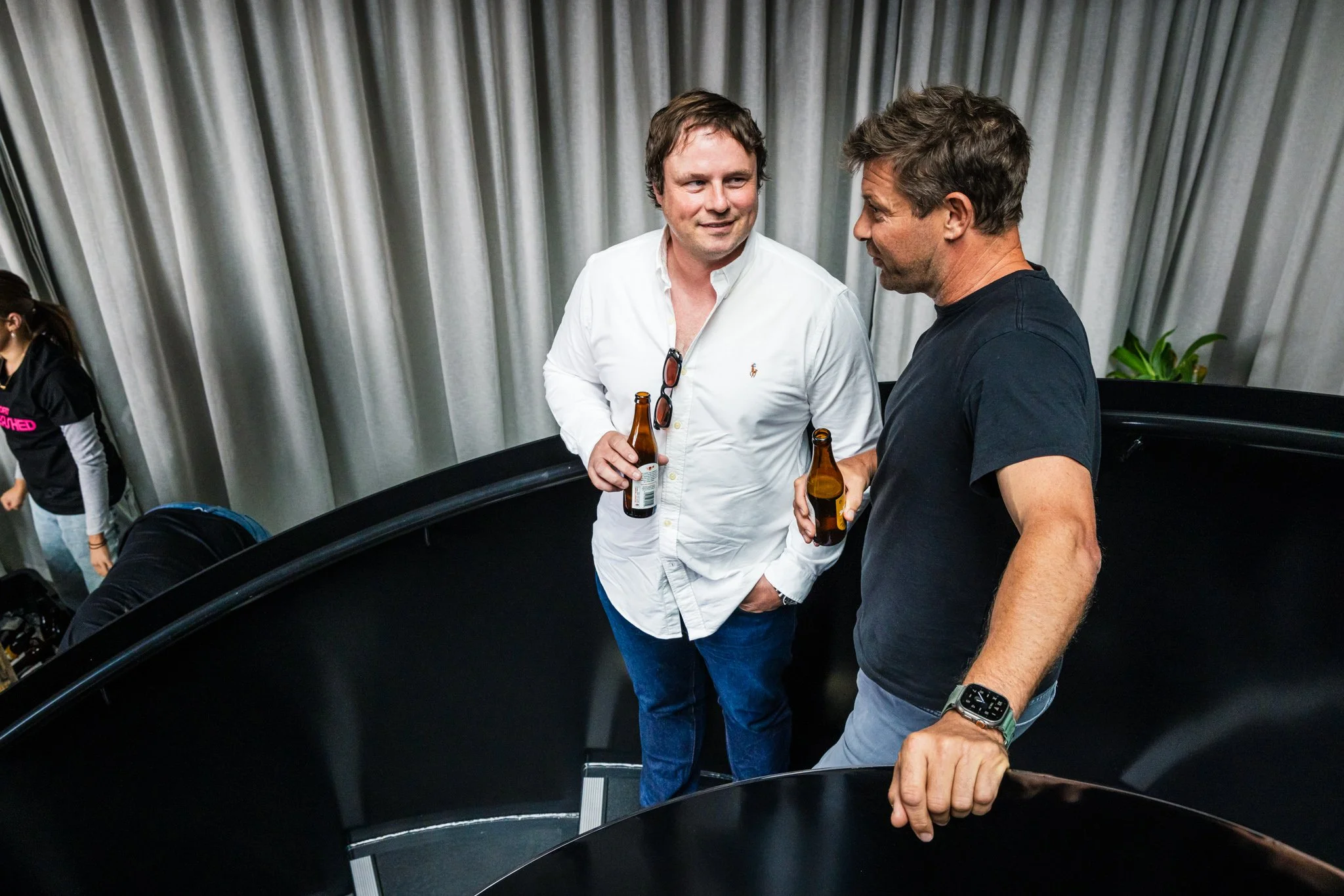 Two men are standing on a staircase, talking and holding beer bottles. One is in a white shirt, the other in a black shirt. The background has grey curtains and a potted plant.
