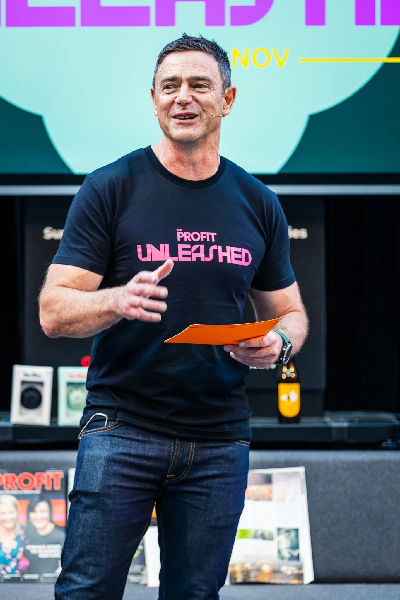 Man speaking at an event, wearing a black t-shirt with pink text that reads 'The Profit UNLEASHED', holding a folder, with a colorful presentation slide in the background.