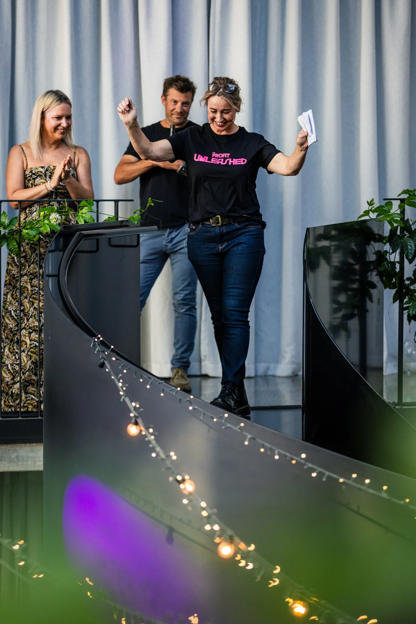 A woman with glasses on her head, wearing a black T-shirt that says 'Profit Unleashed,' walking down a curved corridor with her arms raised in celebration, while two people look on and smile in the background.