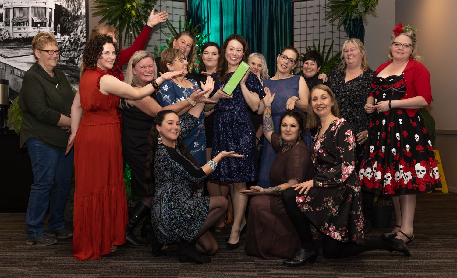 Group of women celebrating at an event, some women are holding a green award, with festive attire and decorations in the background.