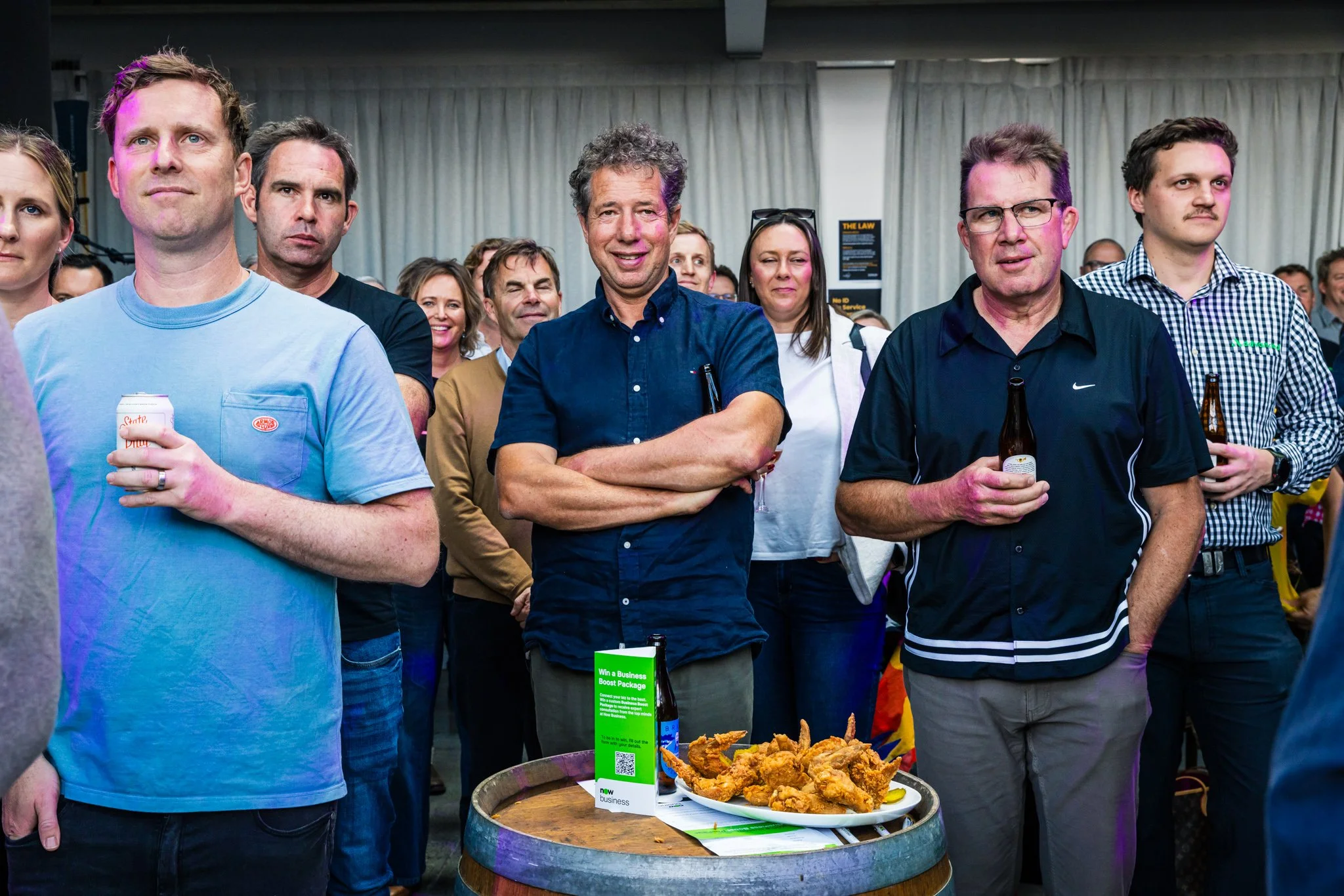 Group of people at a social event, some holding drinks, with a plate of fried chicken on a small barrel in the foreground.