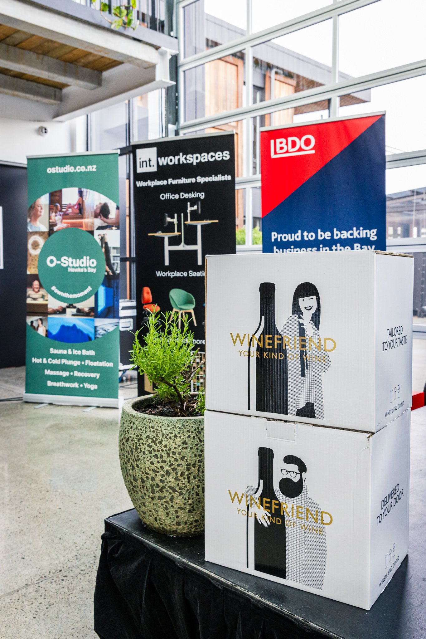 Two stacked white boxes with black and gold illustrations of a man and a woman, labeled "WINEFRIEND" and "Your kind of wine." The boxes are on a black table next to a potted plant. In the background, three vertical banners advertise a workspace furni