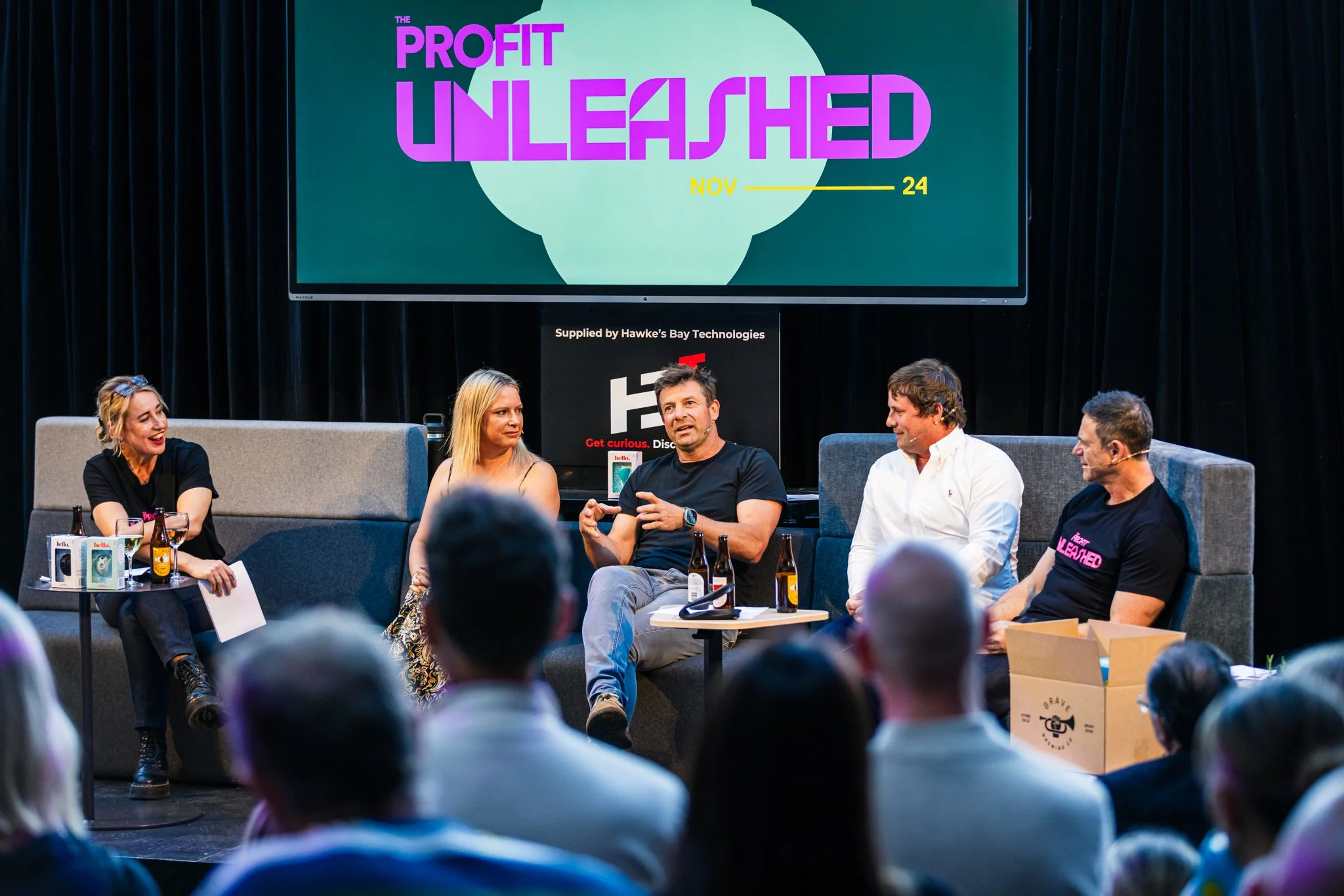 A panel discussion in a dark theater with five people sitting on a stage. Behind them, a large screen displays 'The Profit Unleashed' and the date November 24. Four of the panelists are men and one is woman, engaged in conversation with audience memb