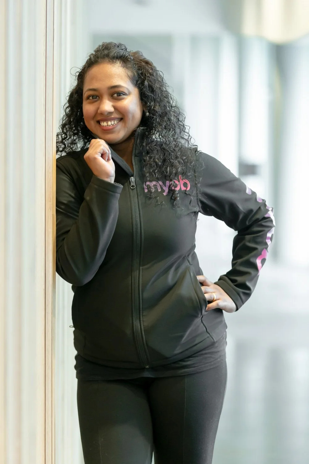 A woman with curly black hair smiling and posing indoors, wearing a black zip-up jacket and black pants.