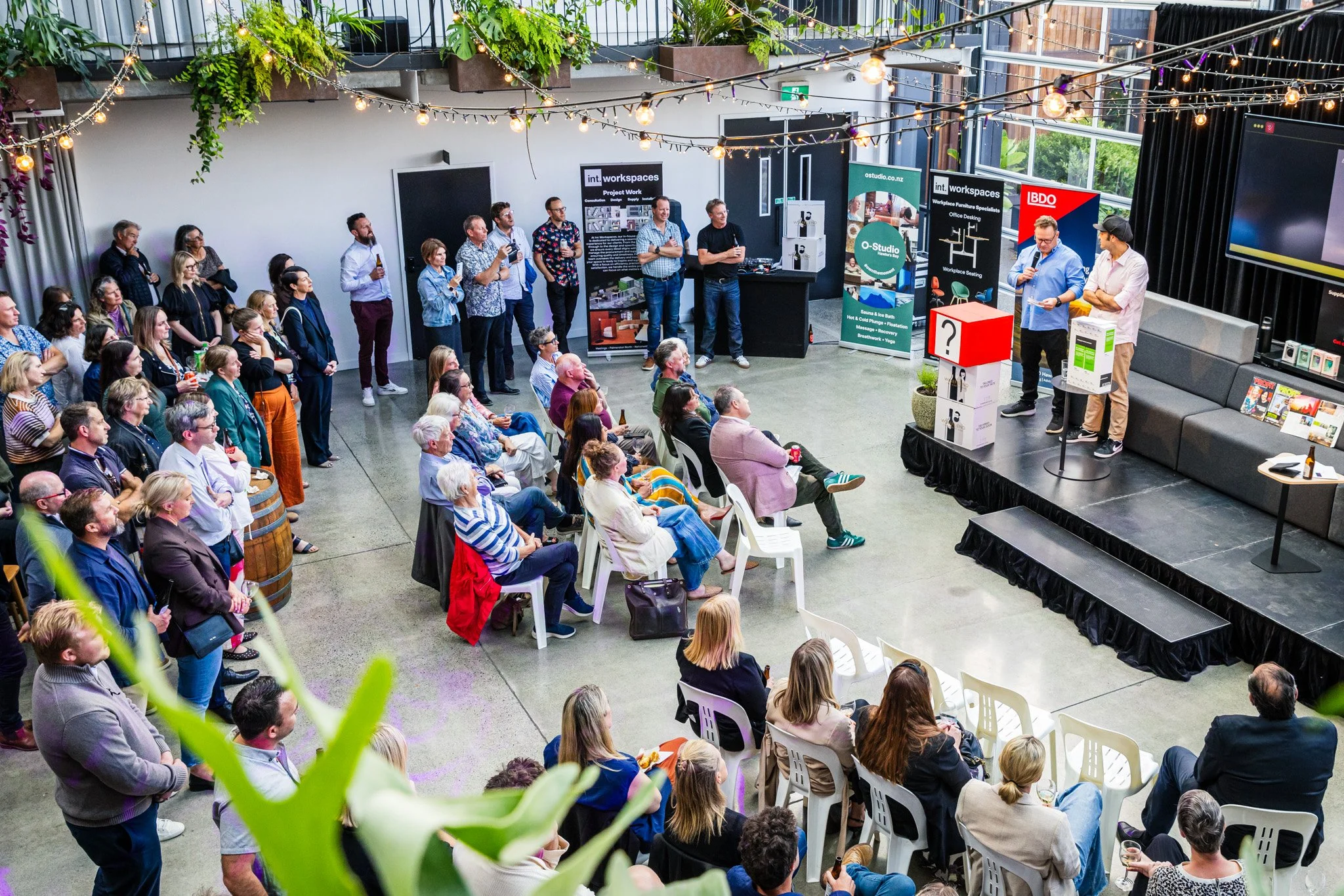 Indoor event with people gathered around a stage, where two men are speaking or presenting. The audience includes seated and standing individuals, with some taking notes or listening attentively. The space has hanging string lights, banner advertisem