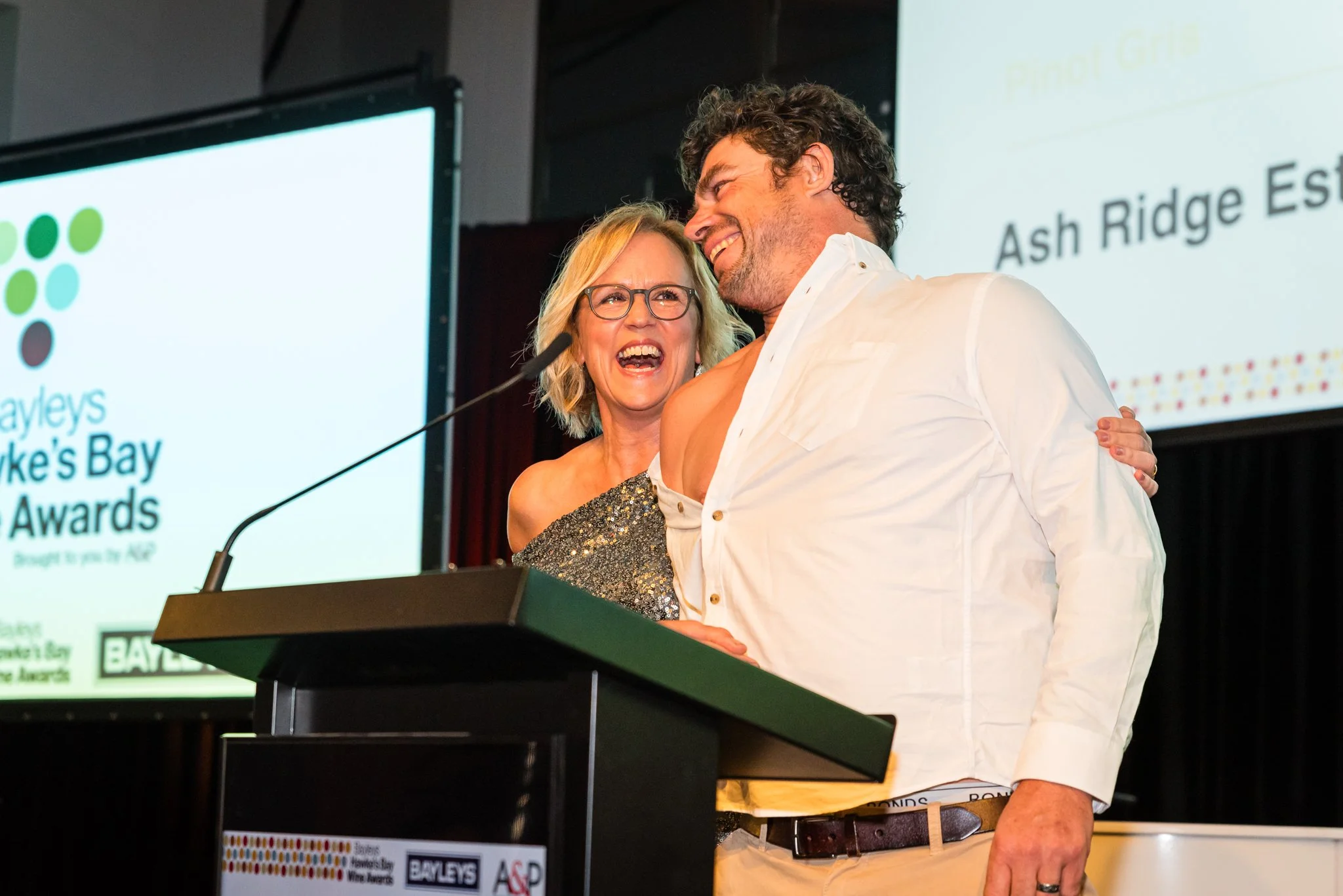 A woman and a man are smiling and hugging at an awards event, with a podium in front of them and a presentation screen behind.