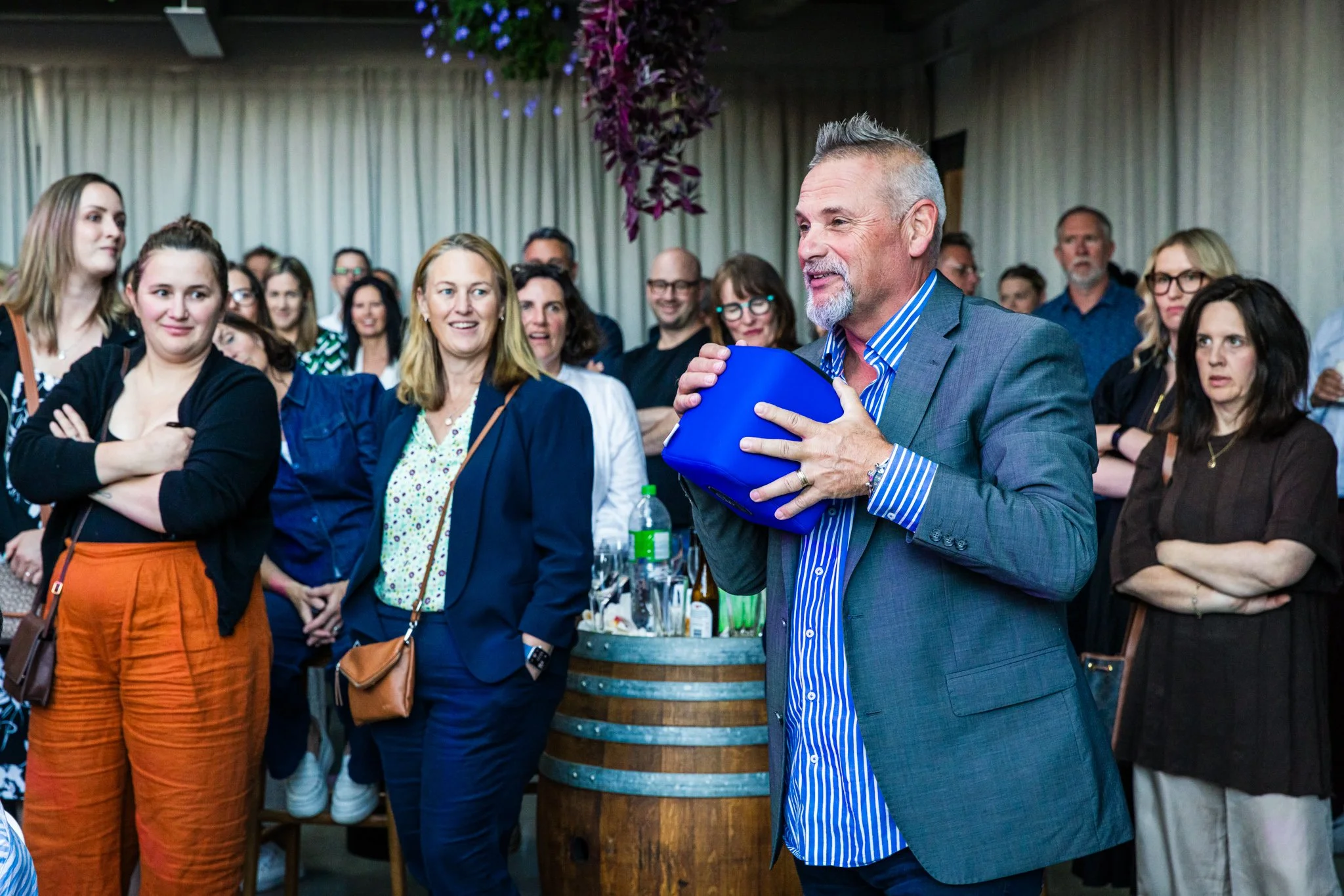 A man in a gray suit holding a blue box, speaking to a group of diverse people in an indoor event space. The audience looks attentive and engaged.