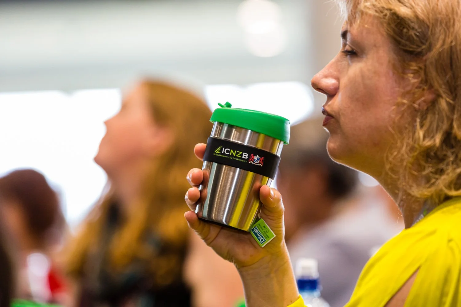A woman with curly blonde hair holding a stainless steel tumbler with a green lid, sitting in a crowded room.