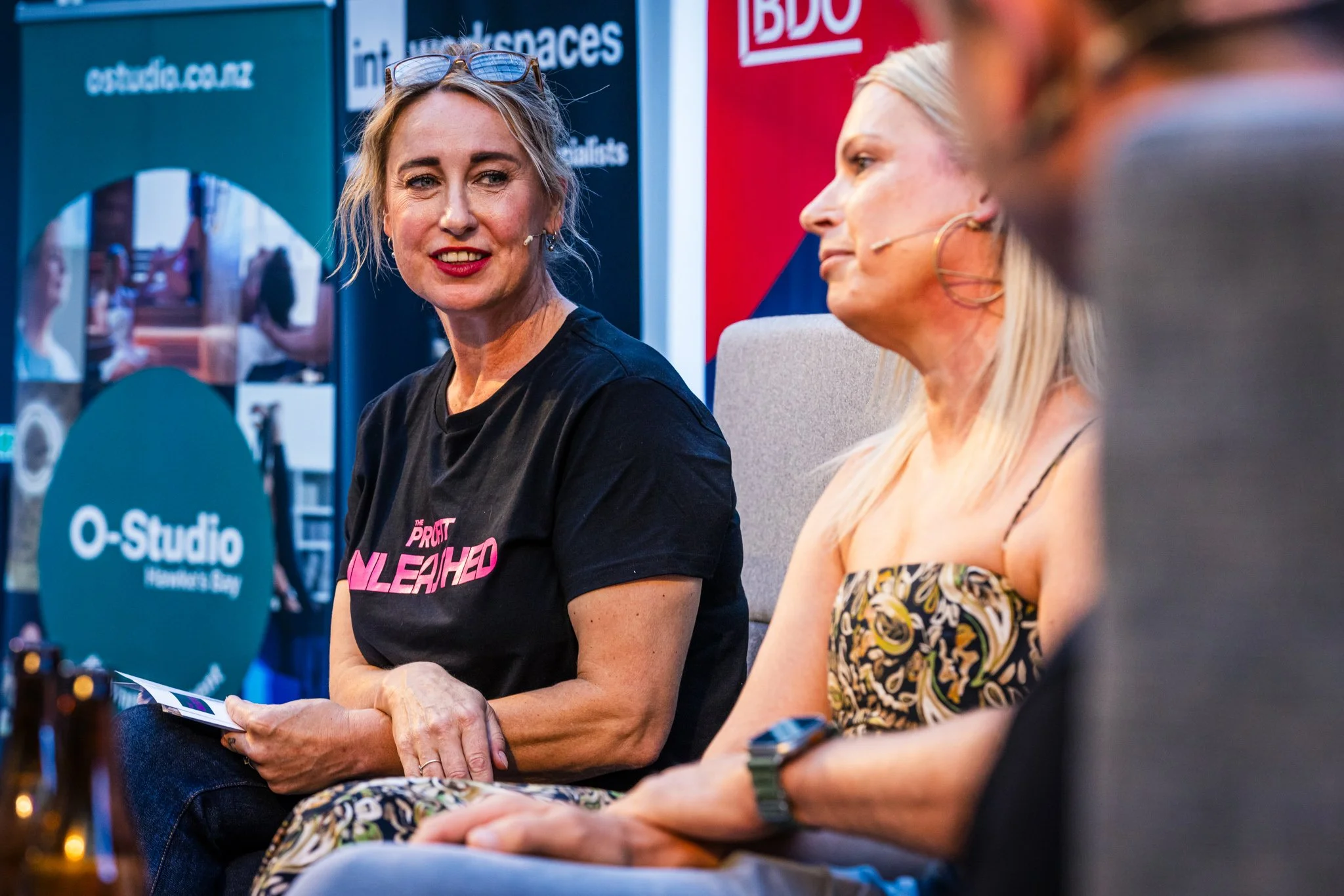 Two women seated on a stage participating in a discussion, with microphones attached to their faces. The woman on the left, with blonde hair, is wearing a black t-shirt with pink text. The woman on the right has long blonde hair and is wearing a patt