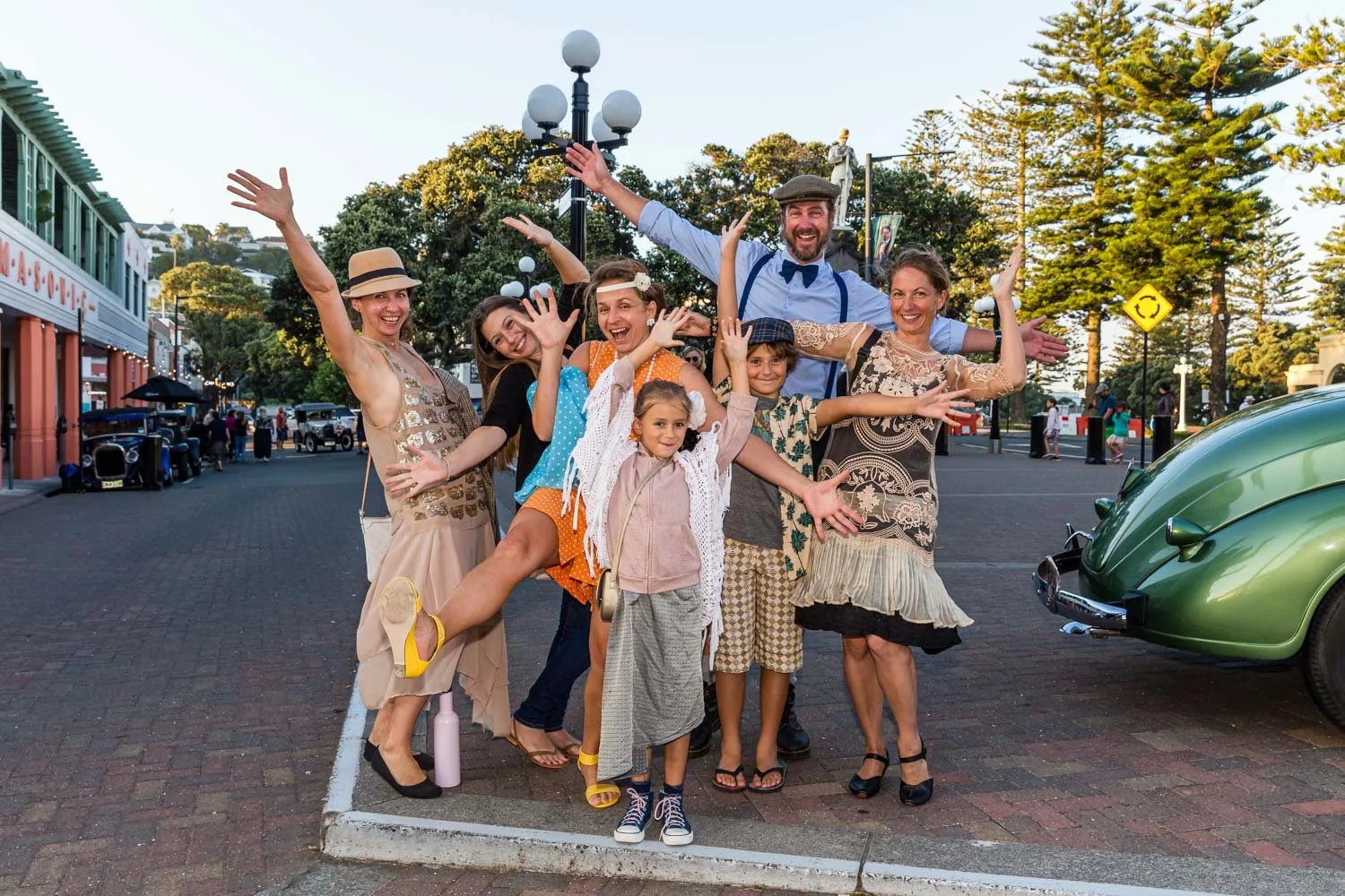 A joyful group of seven people, including children and adults, smiling and posing with their arms raised on a city street lined with trees, vintage cars, and shops, during daytime.