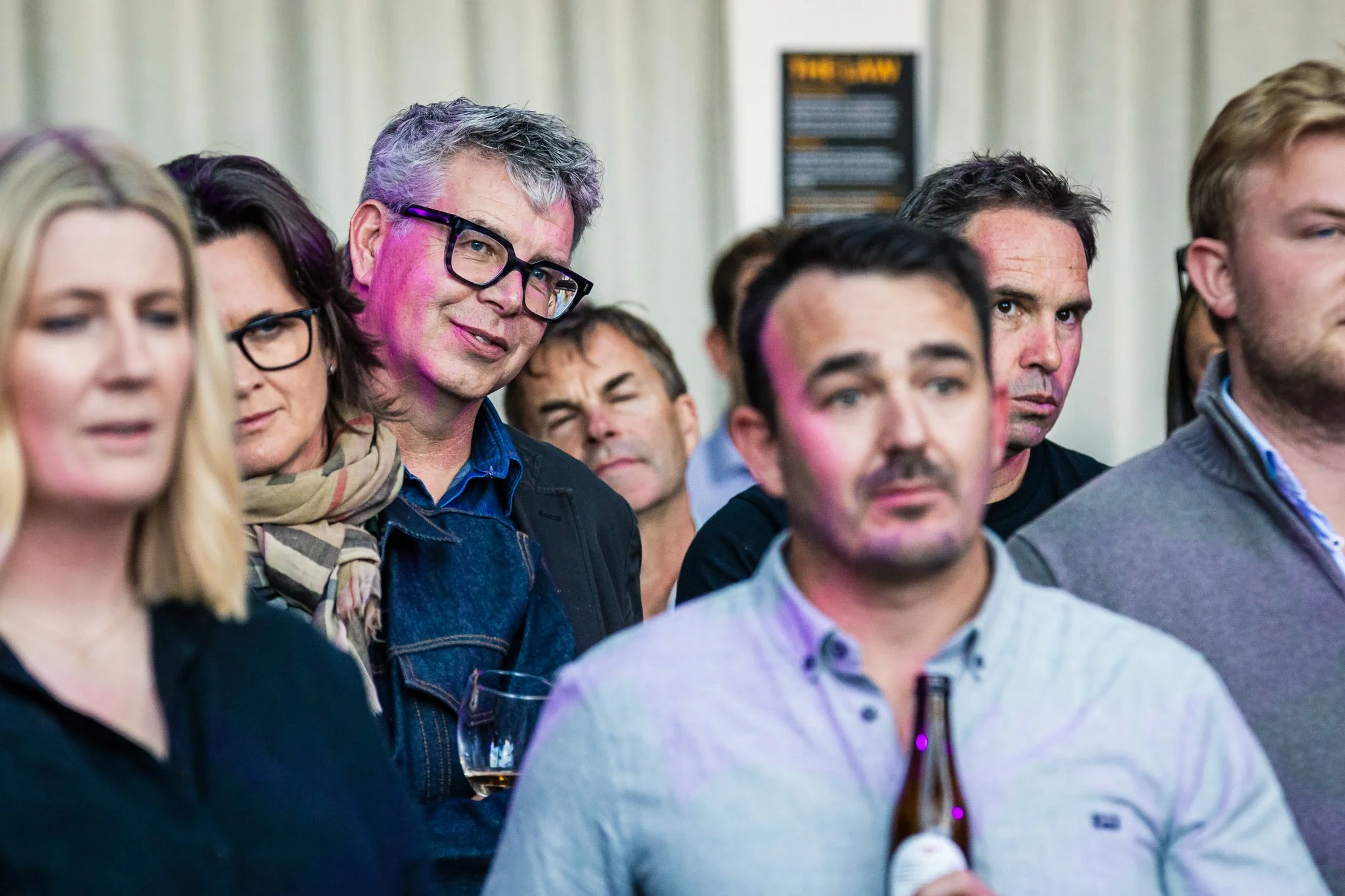 A group of people attending a social event or gathering, with some looking serious and others appearing contemplative; one person is holding a beer bottle.